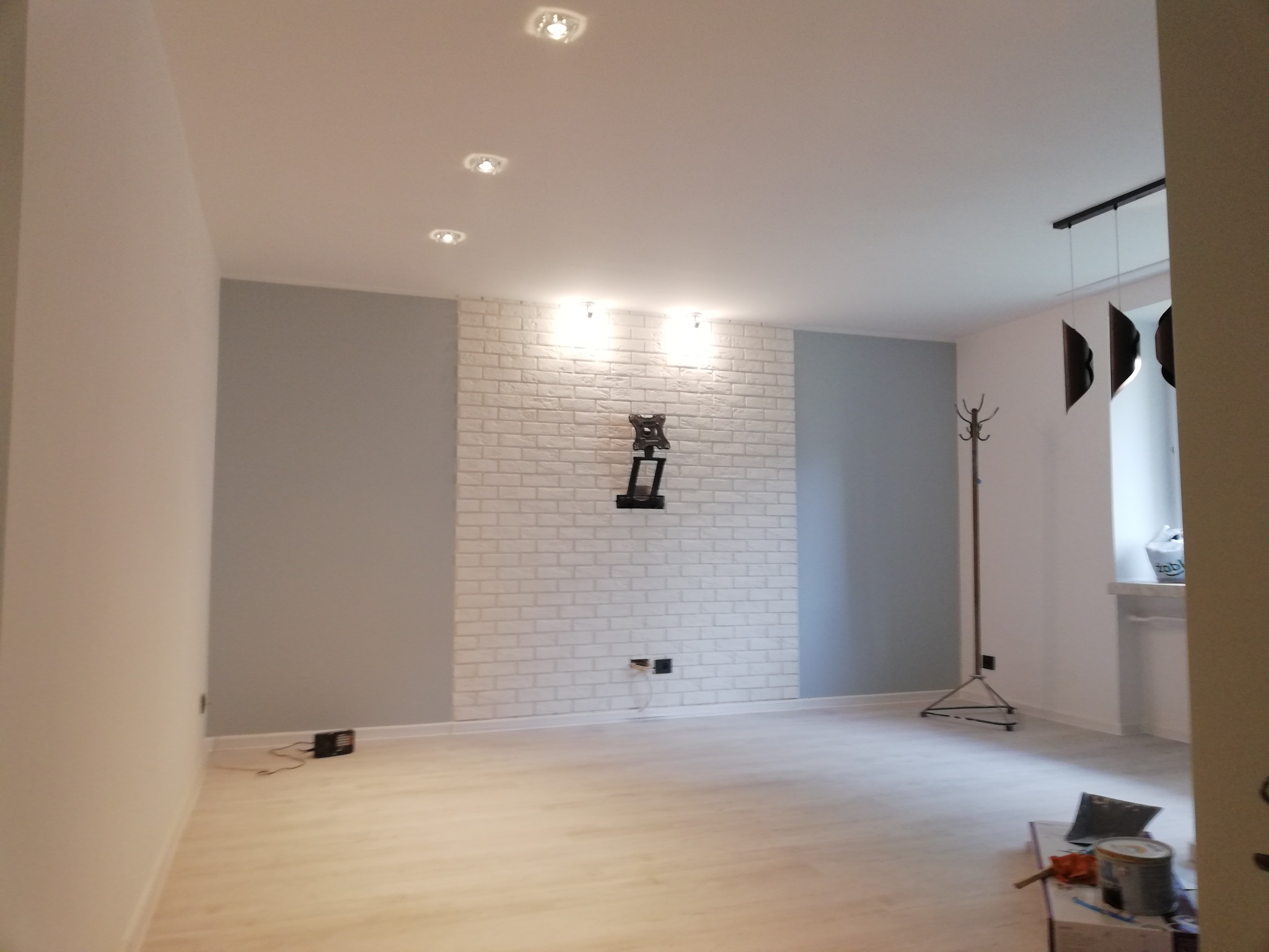 Jasne wnętrze pokoju po odświeżeniu: biała cegła na ścianie, szare ściany boczne, jasna podłoga z widocznymi narzędziami i puszką farby, uchwyt na telewizor na ścianie.