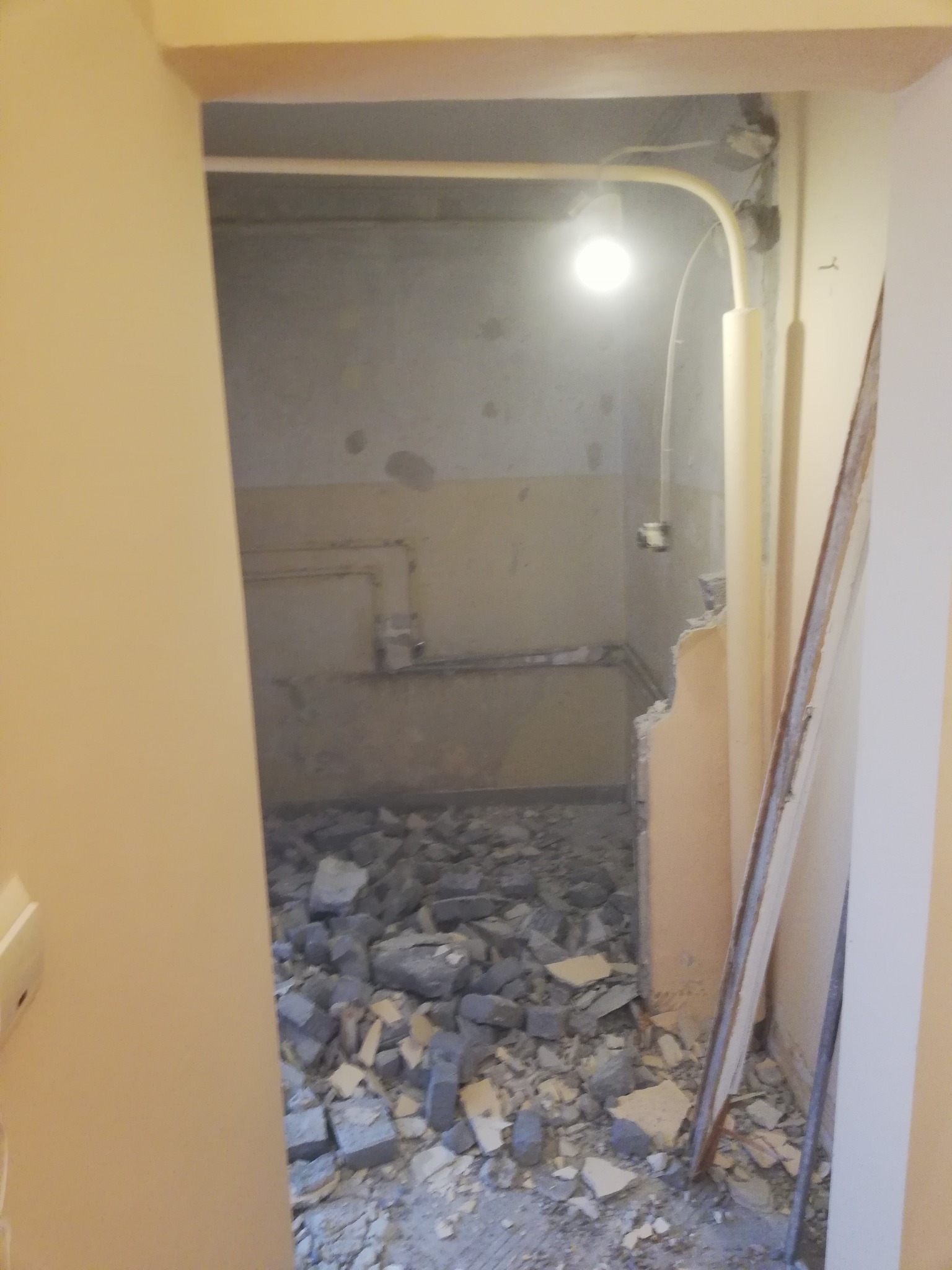 Wnętrze w trakcie demontażu, widoczne gruz i fragmenty ściany, surowa żarówka oświetla przestrzeń, widok z korytarza.