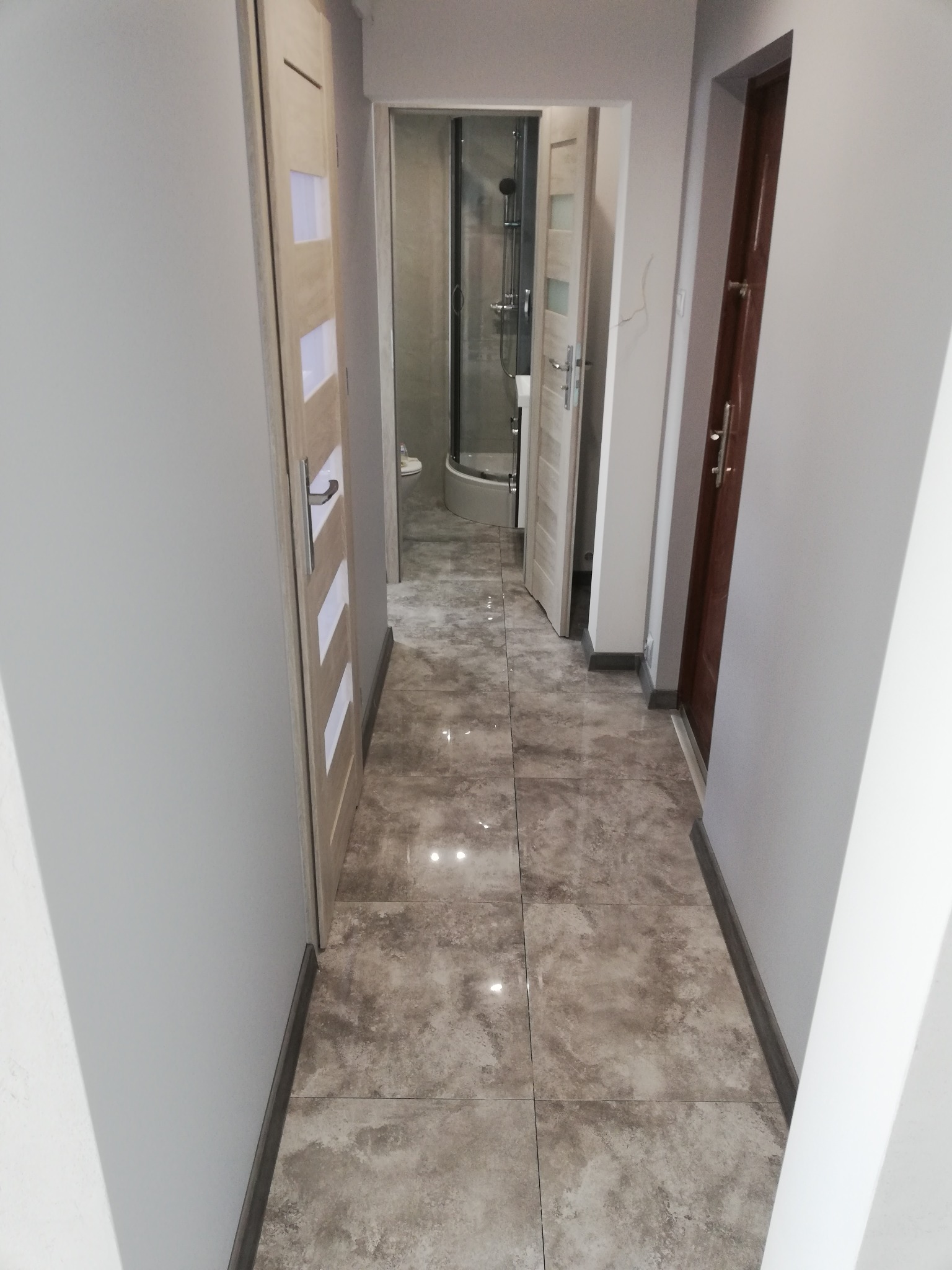 Korytarz z szarymi ścianami, płytkami imitującymi marmur na podłodze, widok na łazienkę z kabiną prysznicową i dwoma parami drzwi po bokach, jedne z drewnianymi wstawkami.
