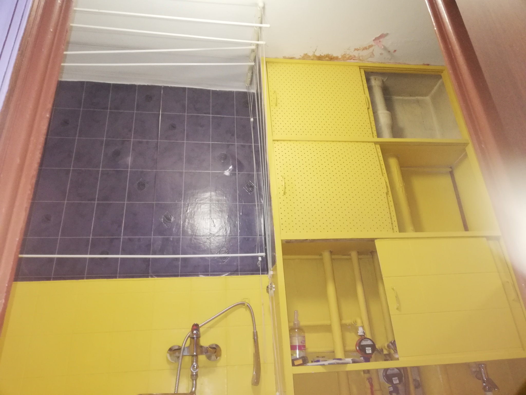 Żółta łazienka z ciemnymi płytkami, widoczna bateria umywalkowa, suszarka na pranie pod sufitem i szafki z perforowanymi drzwiczkami, zdjęcie z dołu do góry.