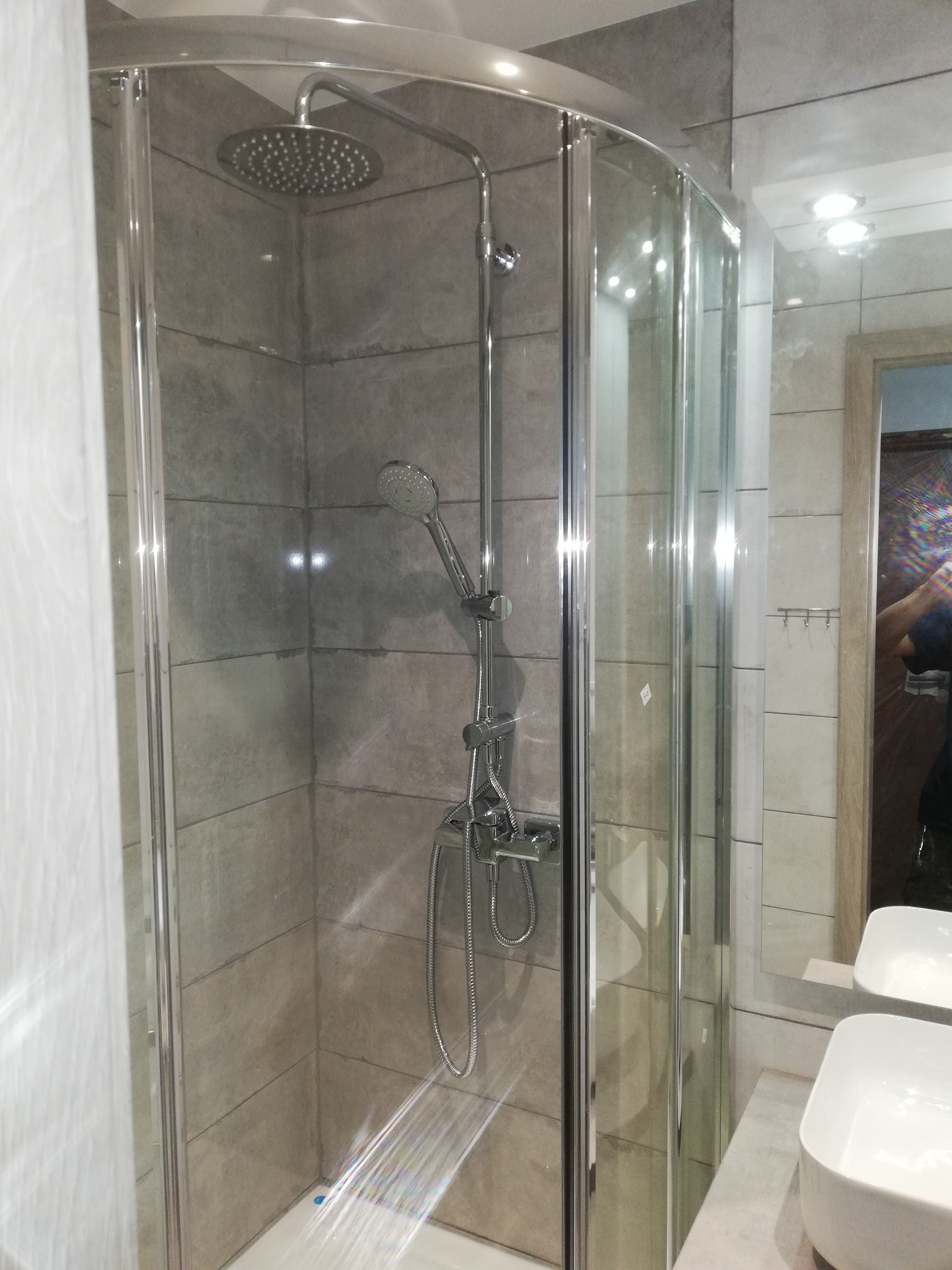 Nowoczesna kabina prysznicowa z deszczownicą i baterią termostatyczną, wyłożona szarymi płytkami imitującymi kamień, z widocznym fragmentem umywalki i lustra.