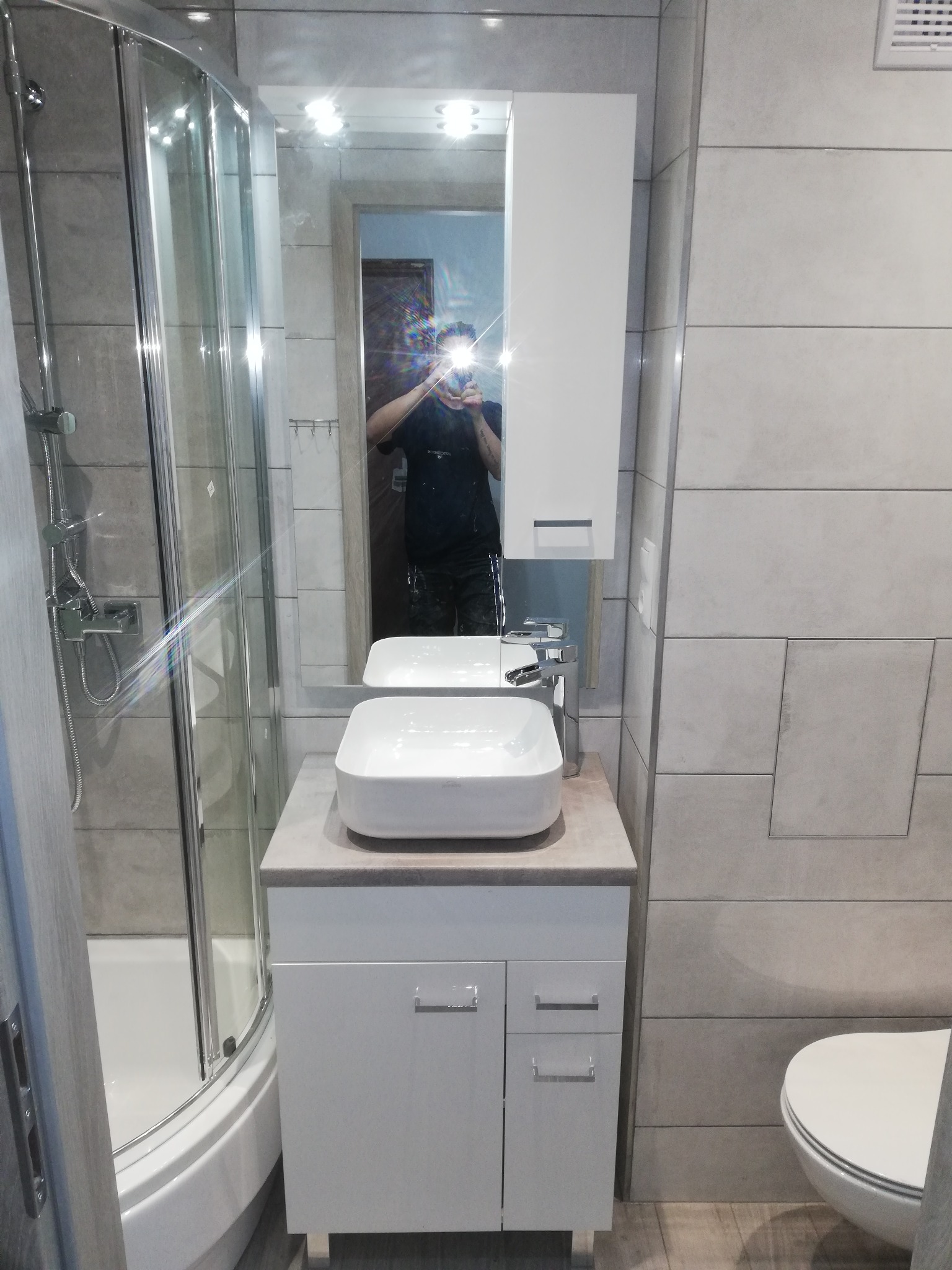 Wykończona łazienka z kabiną prysznicową, umywalką nablatową na szafce z szufladami, lustrem z szafką i ukrytą klapą rewizyjną w płytkach.