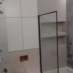 Nowoczesna łazienka z białą zabudową meblową, toaletą podwieszaną, parawanem nawannowym z czarną ramą i oświetleniem LED pod wanną.