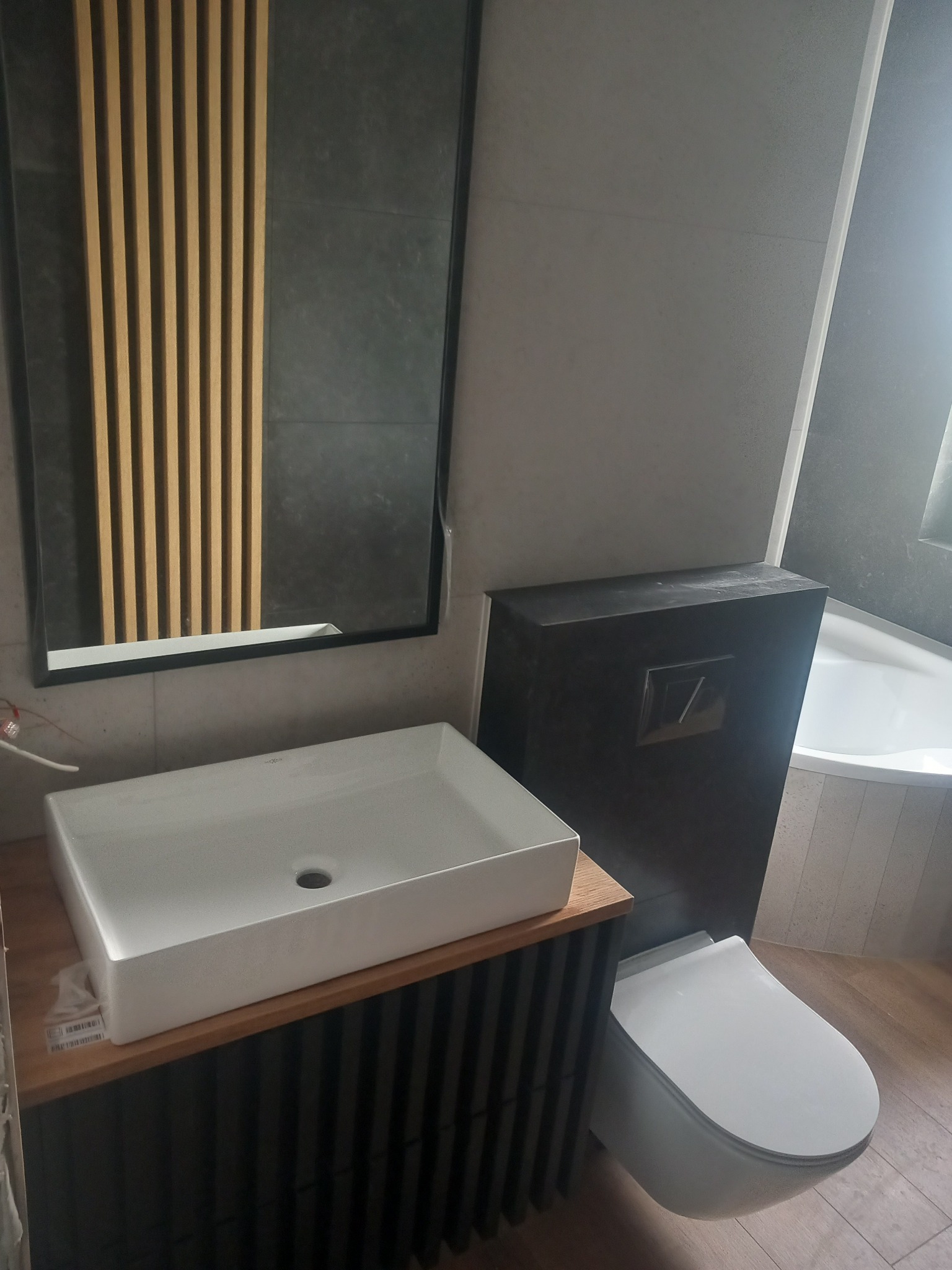 Nowoczesna łazienka z prostokątną umywalką nablatową, lustrem z drewnianymi lamelami, podwieszanym WC i wanną w tle.