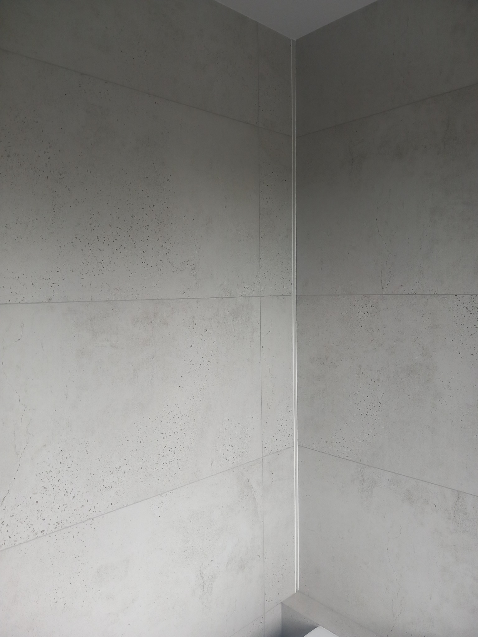 Ściana łazienki wyłożona szarymi płytami imitującymi beton, z widocznymi fugami i narożnikiem, minimalistyczny design.