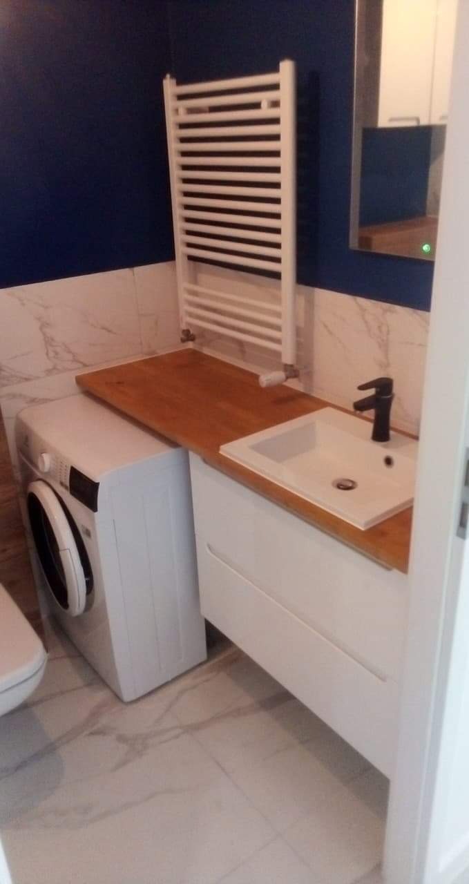 Nowoczesna łazienka z pralką pod blatem, biała umywalka z czarną baterią, grzejnik drabinkowy na ścianie w kolorze granatowym, marmurowe płytki podłogowe i ścienne.