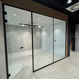 OK Clean Home - Nowoczesne biuro z przeszklonymi ścianami działowymi i otwartymi drzwiami, ukazujące puste, odświeżone wnętrze z szarą podłogą i ścianami. Widoczny system wentylacji sufitowej.