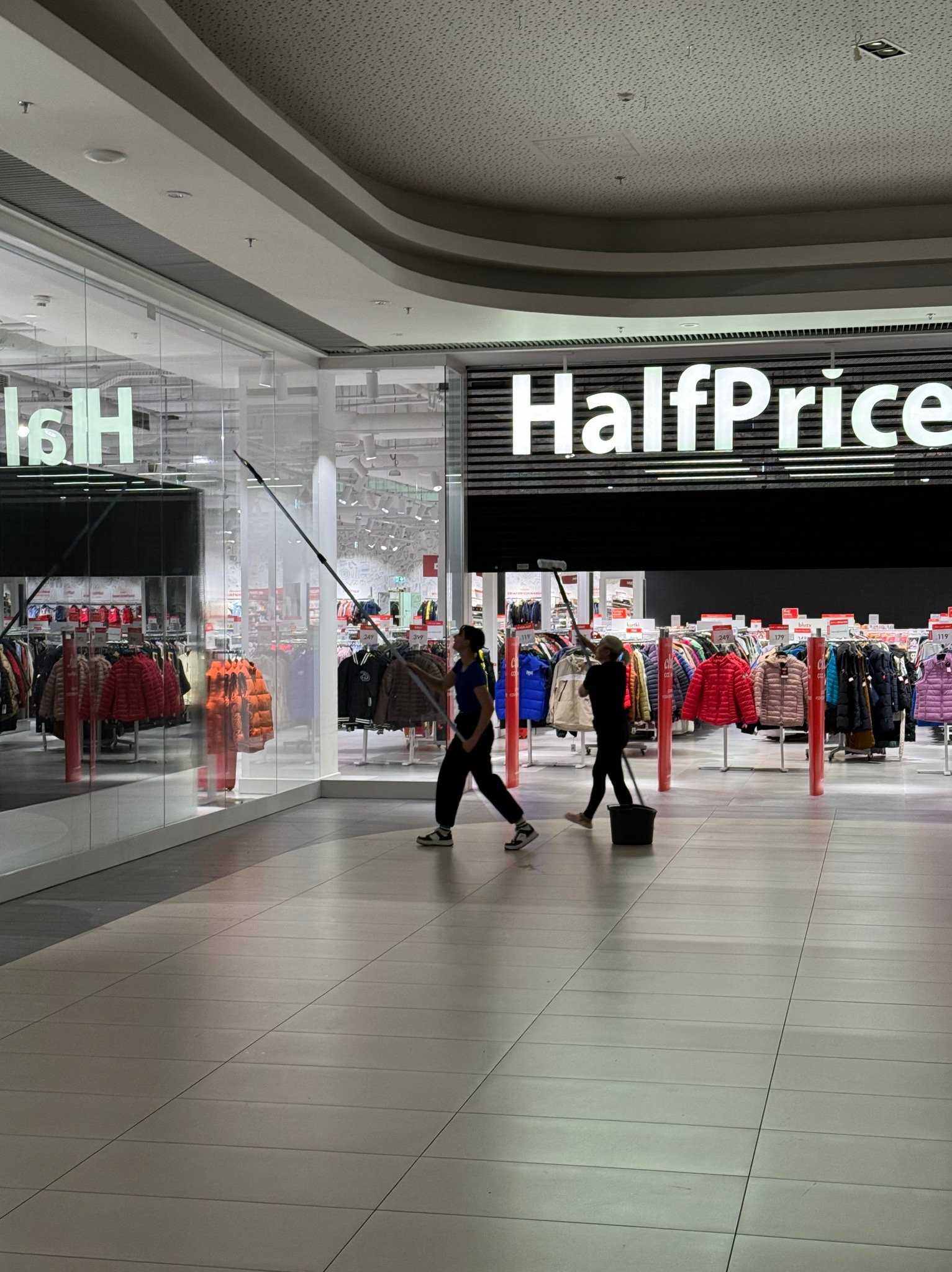 Dwie osoby myją wysokie okna wystawowe sklepu HalfPrice w galerii handlowej za pomocą długich kijów i wiadra.