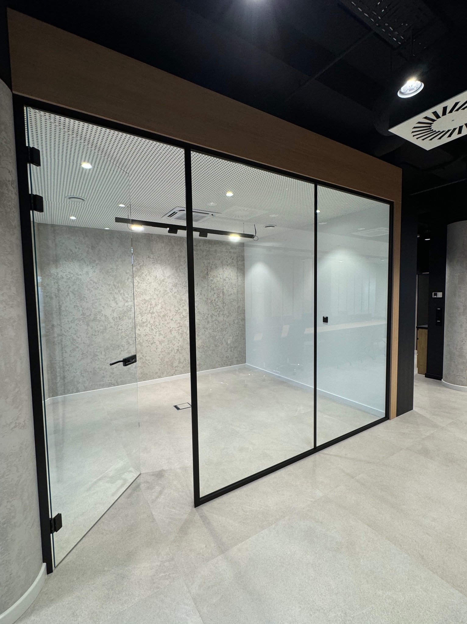 Nowoczesne biuro z przeszklonymi ścianami działowymi i otwartymi drzwiami, ukazujące puste, odświeżone wnętrze z szarą podłogą i ścianami. Widoczny system wentylacji sufitowej.