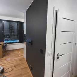 PSP PIOTR GOLEC GITCHATA - Minimalistyczne wnętrze: czarna ściana z uchwytem na telewizor, drewniana podłoga, białe drzwi z poziomymi frezami i czarne rolety w oknie.