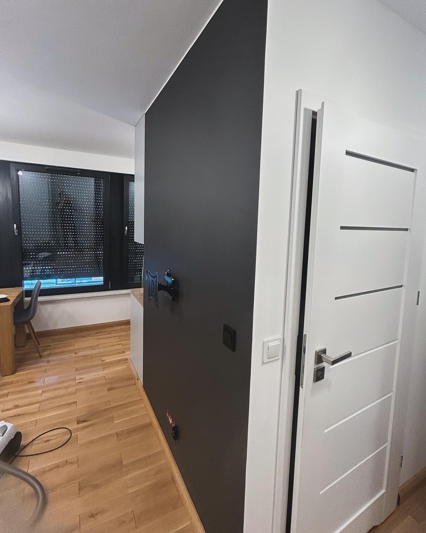 Minimalistyczne wnętrze: czarna ściana z uchwytem na telewizor, drewniana podłoga, białe drzwi z poziomymi frezami i czarne rolety w oknie.
