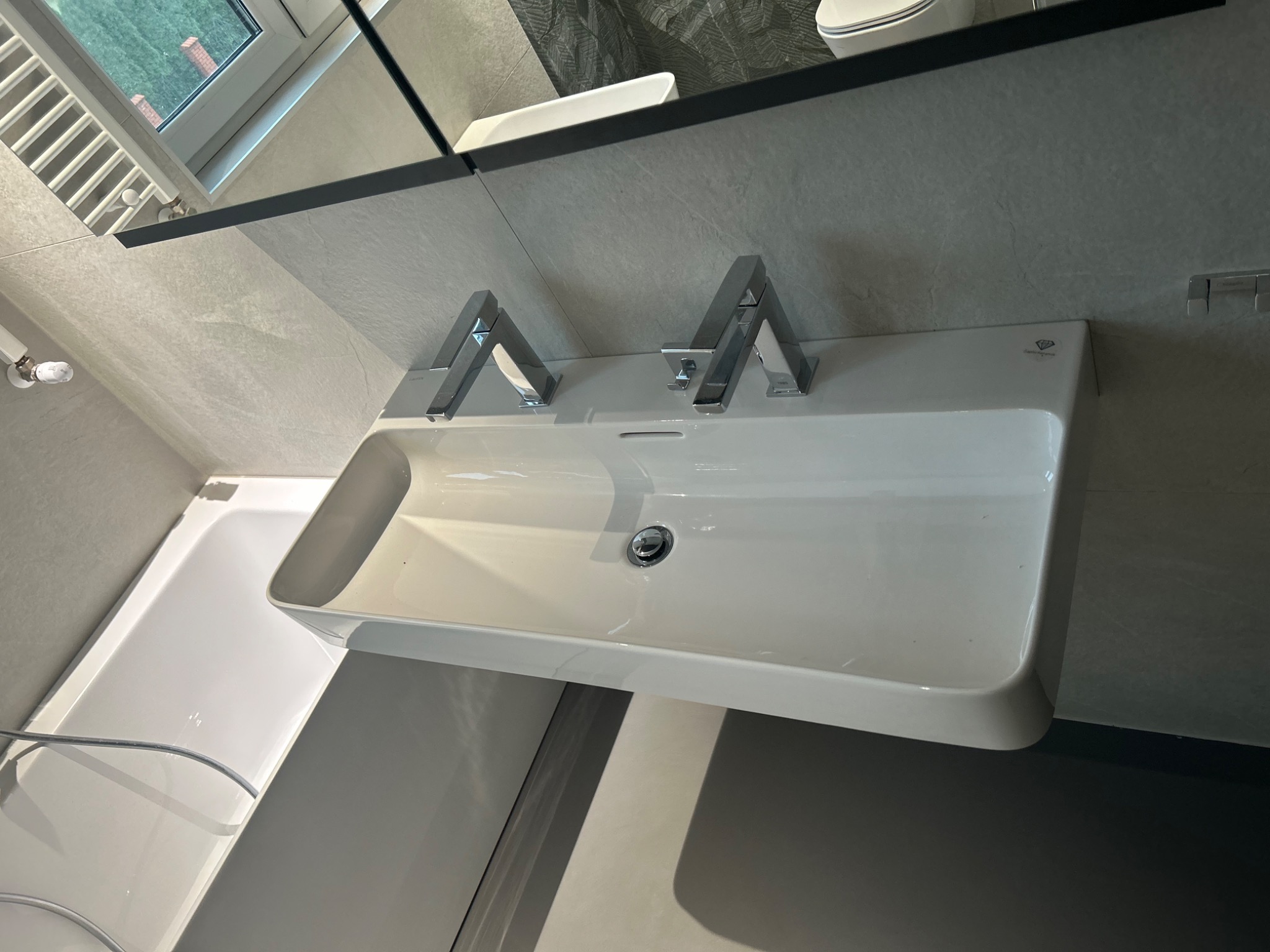 Nowoczesna, podwójna umywalka nablatowa z chromowanymi bateriami, zamontowana na szarej, kamiennej ścianie, z widocznym fragmentem wanny i grzejnika łazienkowego w tle.