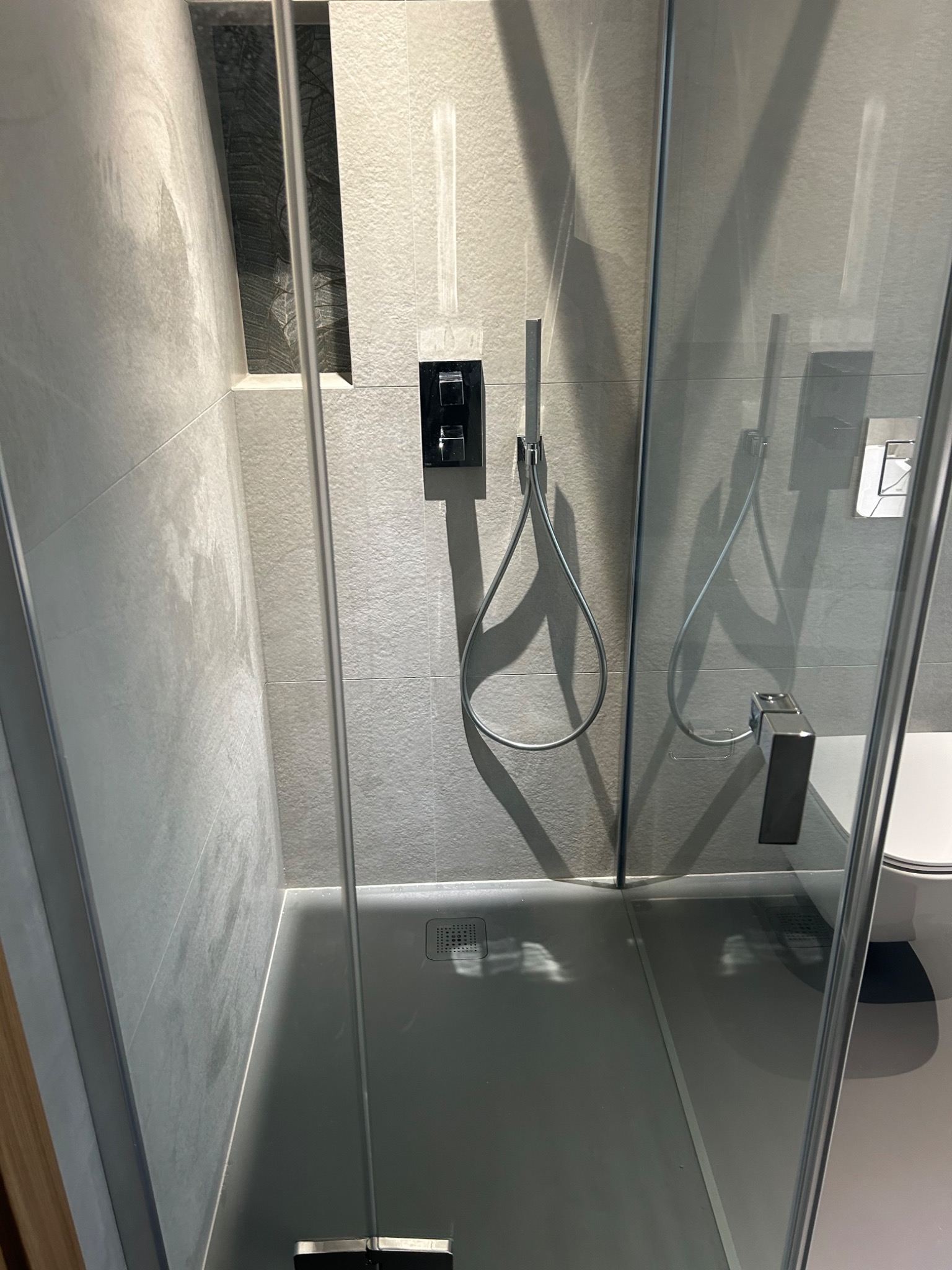 Nowoczesna kabina prysznicowa z szarymi płytkami, deszczownicą i minimalistyczną armaturą, widok z perspektywy wejścia, z widocznym odpływem liniowym.