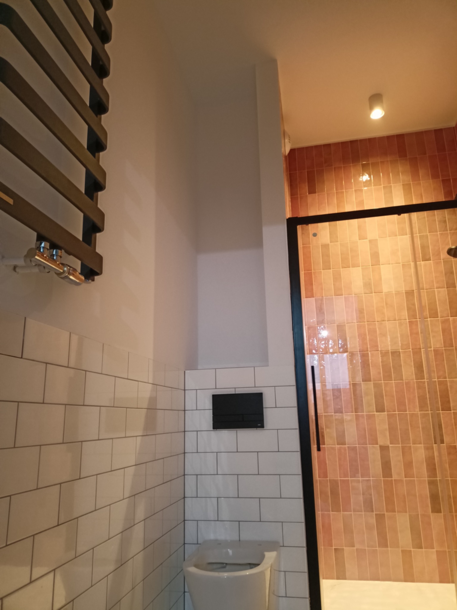 Nowoczesna łazienka z białymi, prostokątnymi płytkami ułożonymi do połowy ściany, grzejnikiem drabinkowym w kolorze ciemnego brązu, wiszącą miską WC i kabiną prysznicową z czarnymi profilami...