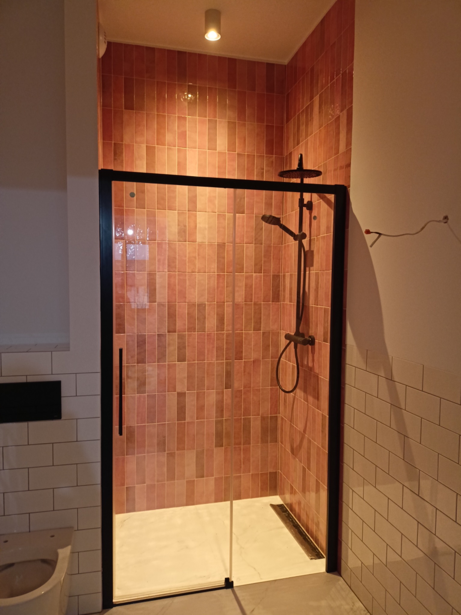 Wykończona kabina prysznicowa typu walk-in z czarną ramą i deszczownicą, obłożona pionowo ułożonymi, wąskimi, różowo-pomarańczowymi płytkami, widoczna częściowo toaleta i płytki ścienne w stylu metro.