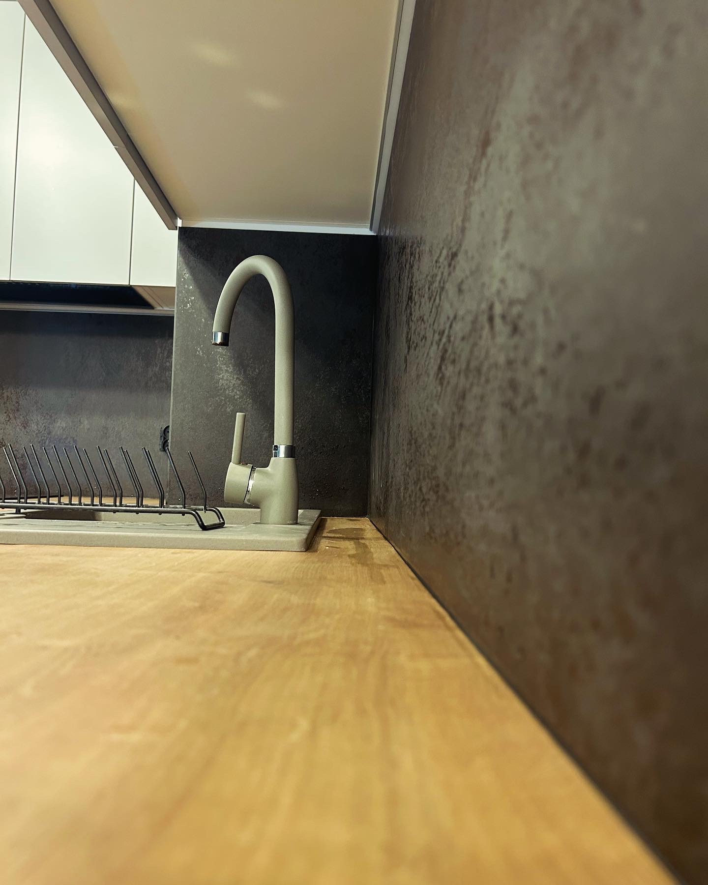 Szary kran z wyciąganą wylewką i ociekaczem na naczynia na blacie z drewnopodobnego laminatu, tło stanowi ściana w kolorze grafitowym z efektem rdzy i białe szafki.