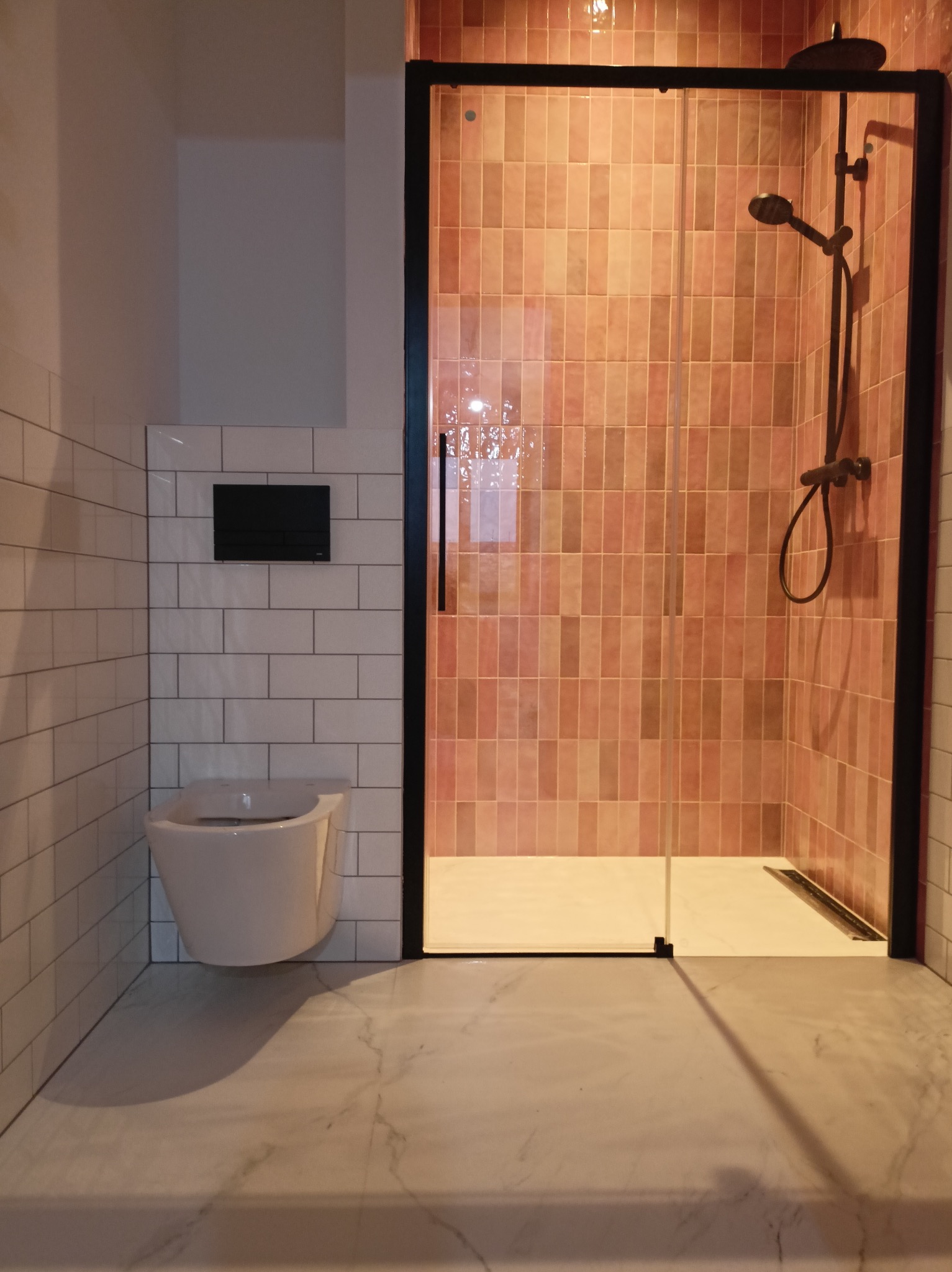 Nowoczesna łazienka z kabiną prysznicową w czarnej ramie i armaturą w kolorze brązu, płytki w odcieniach różu, wisząca toaleta z czarnym panelem i marmurowa podłoga.