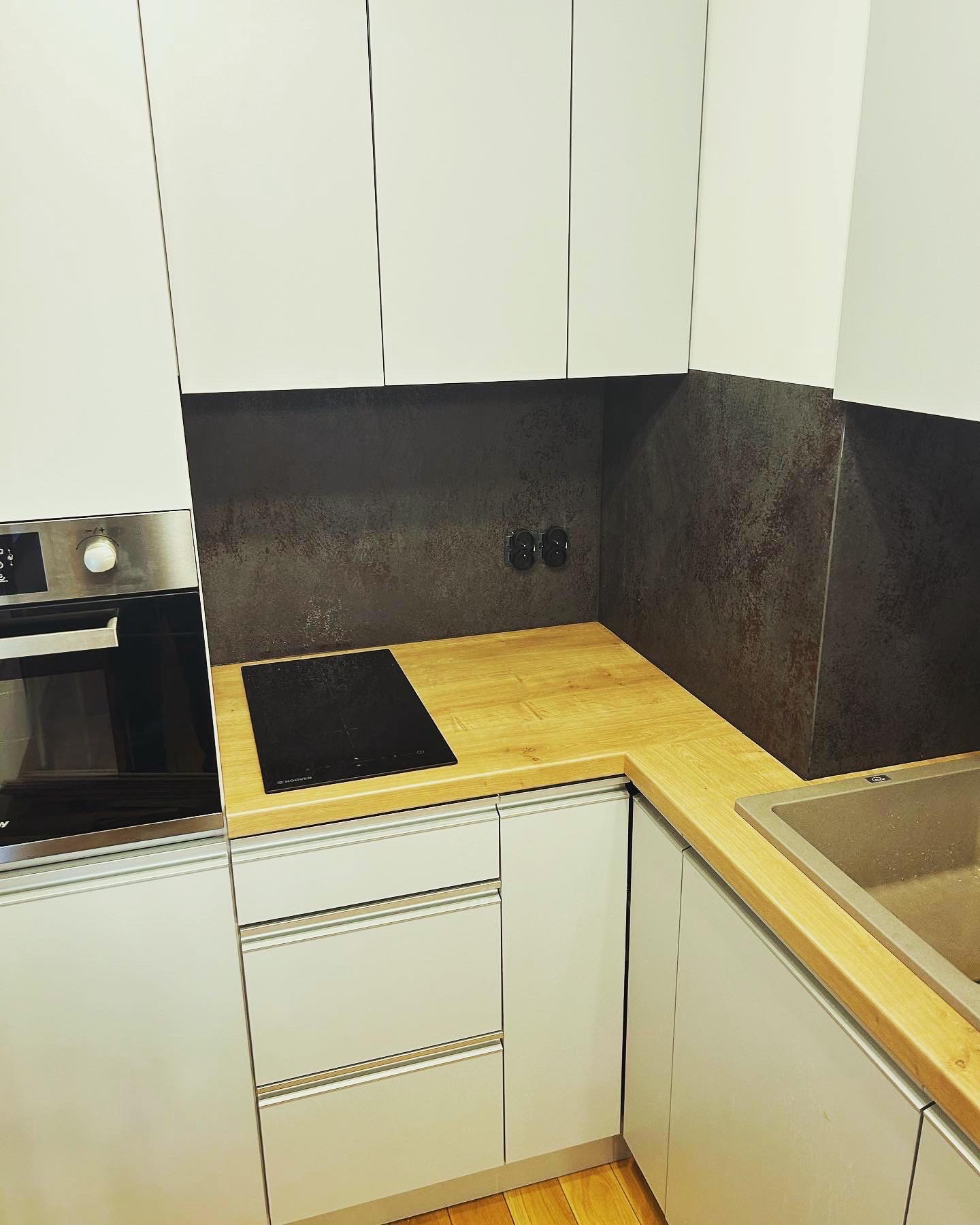 Nowoczesna kuchnia z białymi szafkami, drewnianym blatem, płytą indukcyjną i stalowym piekarnikiem. Ciemna ściana nad blatem z gniazdkami elektrycznymi.