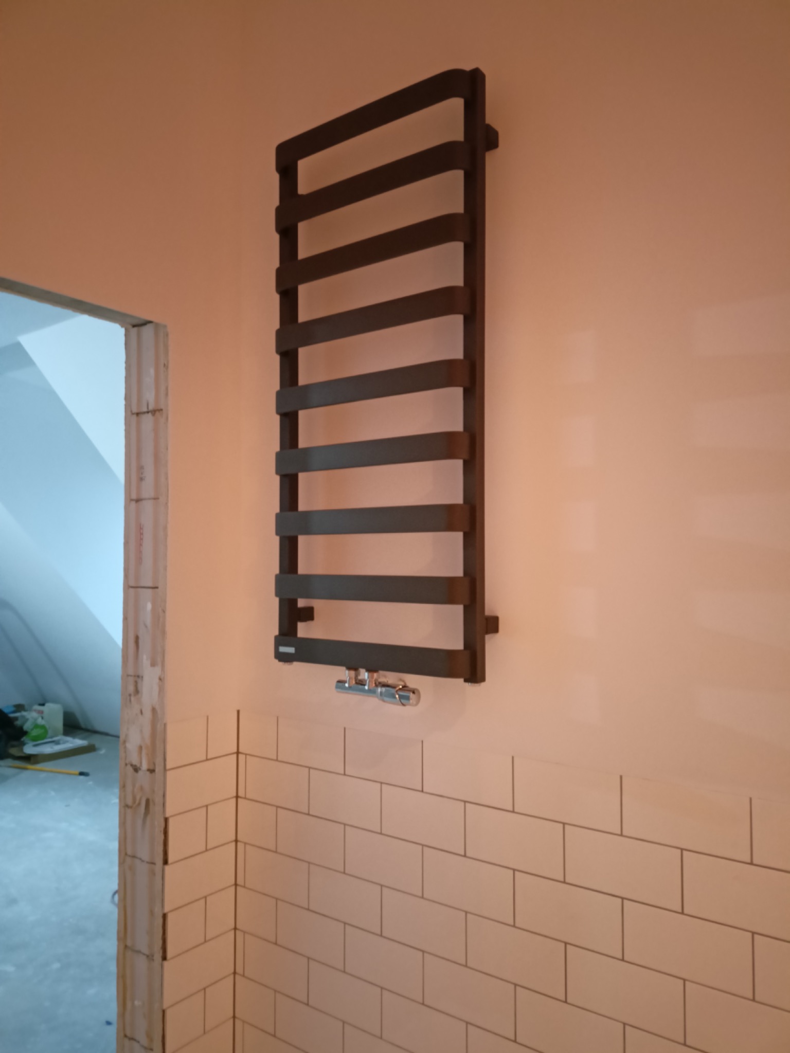 Zamontowany, ciemny grzejnik drabinkowy na ścianie z białymi, prostokątnymi płytkami, widoczny fragment niedokończonego otworu drzwiowego i surowy stan pomieszczenia.