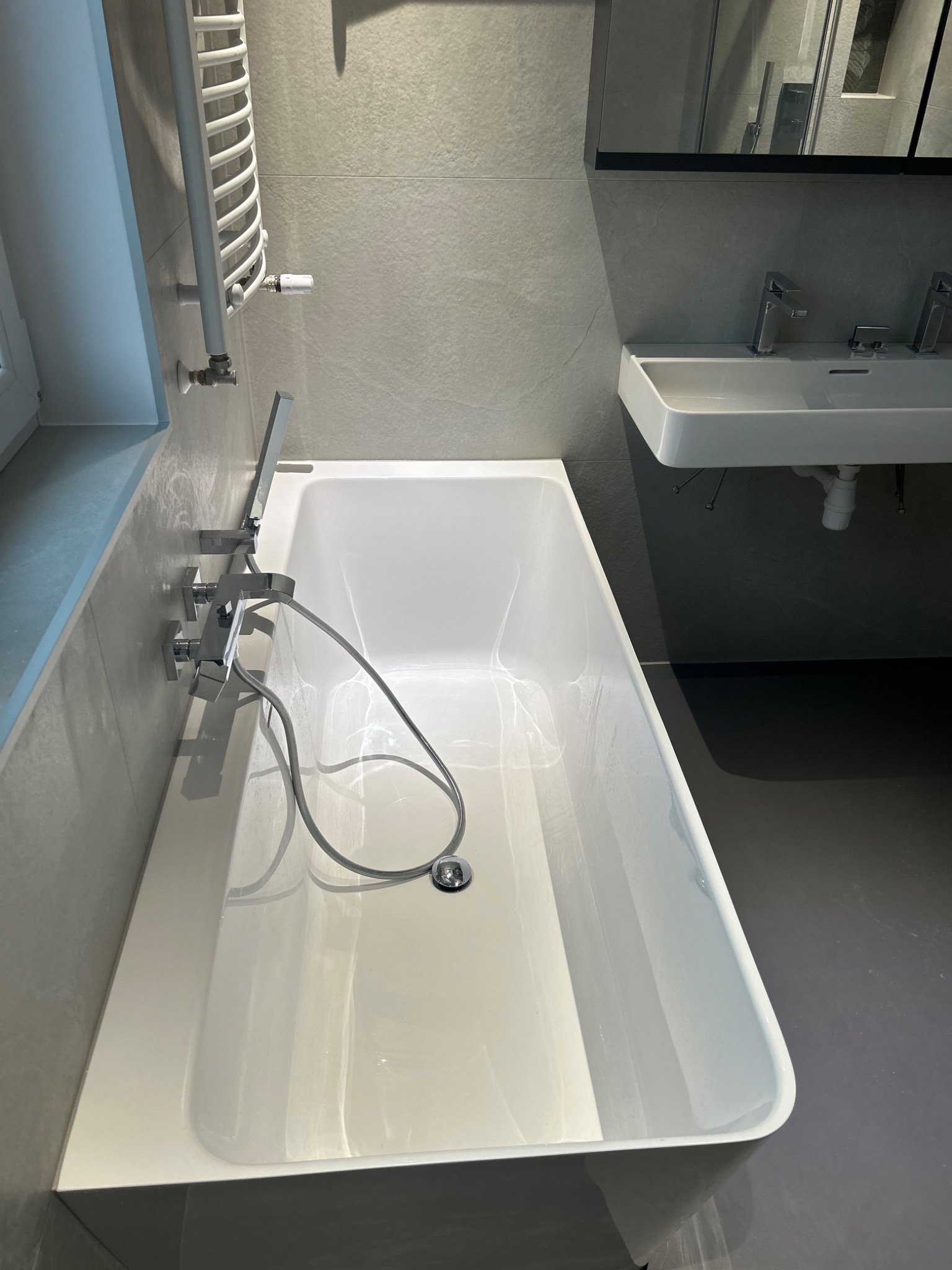 Nowoczesna, biała wanna z chromowaną armaturą i słuchawką prysznicową na tle szarych płytek w łazience. Umywalka z dwoma kranami widoczna w tle.