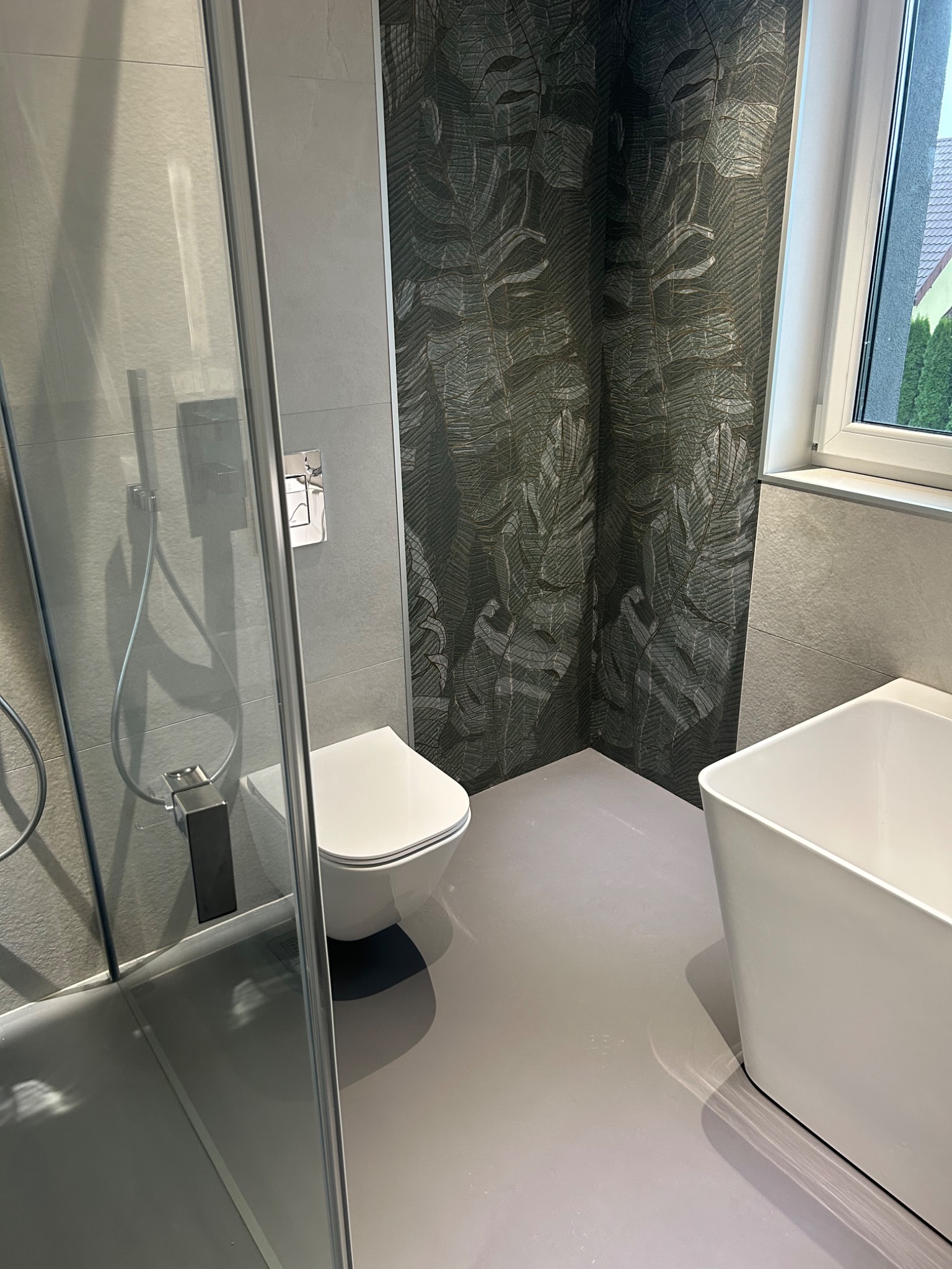 Nowoczesna łazienka z kabiną prysznicową, wiszącą toaletą i wolnostojącą wanną. Ściany wyłożone szarymi płytkami i dekoracyjną tapetą z motywem liści. Szara, jednolita podłoga.
