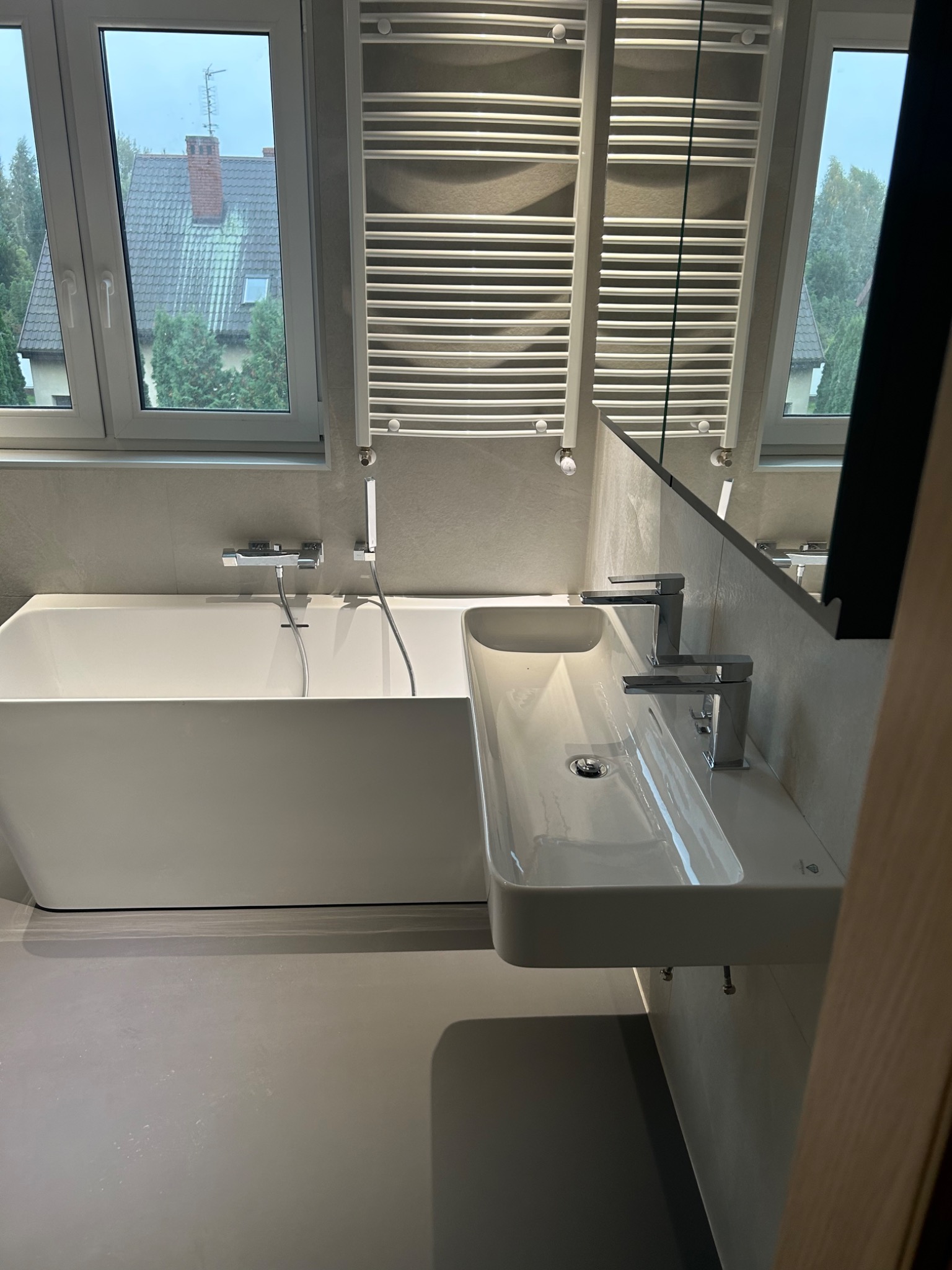 Nowoczesna, jasna łazienka z białą wanną, umywalką nablatową i grzejnikiem drabinkowym, widok przez okno na dom z czerwonym dachem.