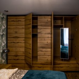 Dobre Quchnie S.C - Drewniana szafa wnękowa z otwartymi drzwiami, z lustrem w środkowej części, widoczna z perspektywy łóżka z zieloną narzutą i poduszkami w liście