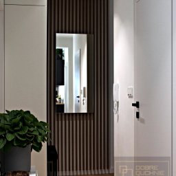 Dobre Quchnie S.C - Nowoczesny przedpokój z lustrem w ramie z drewnianych listewek, białe ściany, drzwi i szafka z rośliną doniczkową, widoczny domofon.