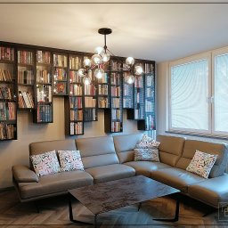 Dobre Quchnie S.C - Salon z szarą sofą narożną, stolikiem kawowym i regałem na książki w zabudowie, oświetlonym nowoczesną lampą wiszącą z żarówkami kulistymi.