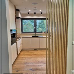 Dobre Quchnie S.C - Jasna kuchnia z drewnianą podłogą i lamelową ścianką działową, widok na okno z zielenią na zewnątrz, nowoczesny design.