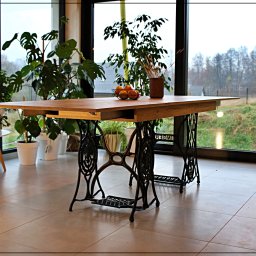 Dobre Quchnie S.C - Drewniany stół z wysuwanym blatem, osadzony na żeliwnych nogach od maszyny do szycia Singer, z misą owoców i wazonem na blacie, na tle okna z widokiem na zewnątrz, otoczony roślinami doniczkowymi.