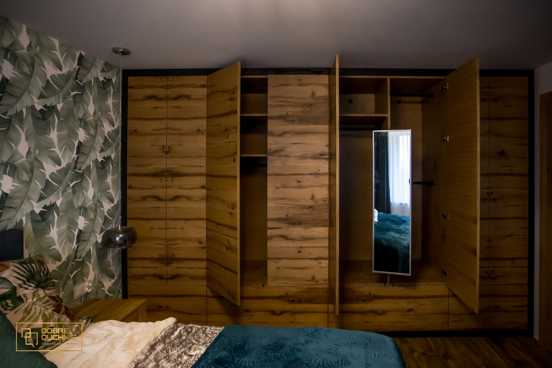 Drewniana szafa wnękowa z otwartymi drzwiami, z lustrem w środkowej części, widoczna z perspektywy łóżka z zieloną narzutą i poduszkami w liście