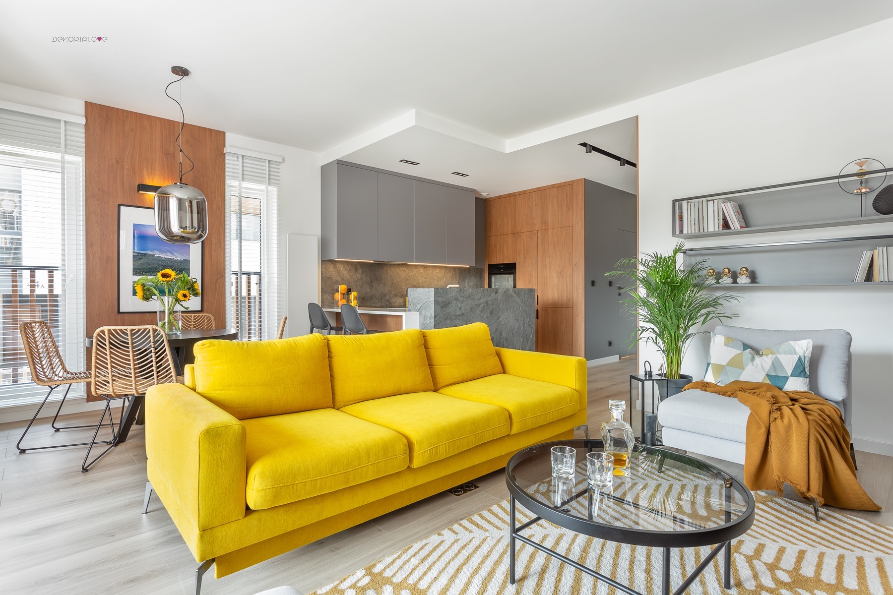 Nowoczesne wnętrze z żółtą sofą, widok na szare szafki kuchenne, drewnianą ścianę i jadalnię z rattanowymi krzesłami, całość dopełniona roślinami i dekoracjami.