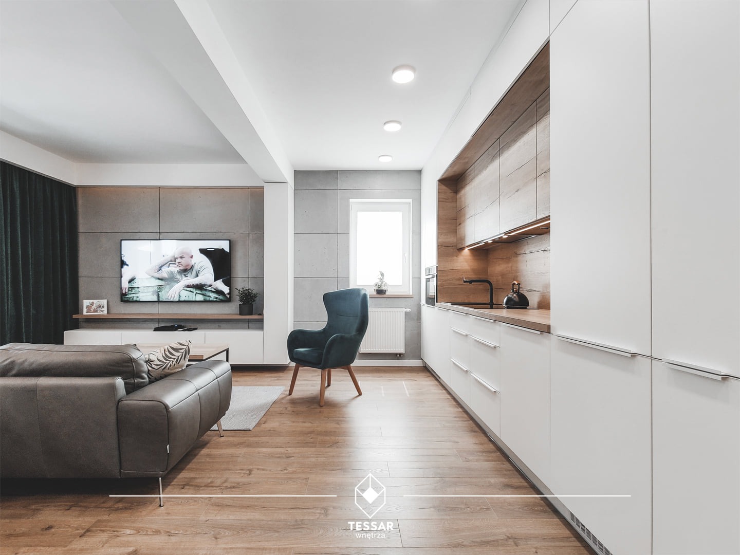 Nowoczesna kuchnia z białymi szafkami i drewnianym blatem, połączona z salonem z szarą sofą i telewizorem na ścianie z betonowych płyt.