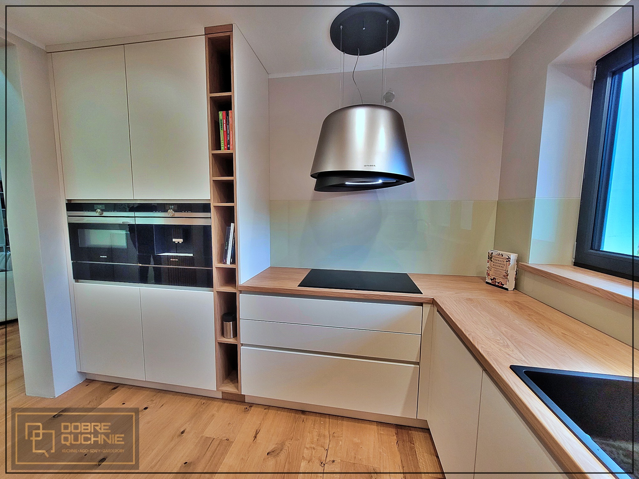 Nowoczesna kuchnia z białymi frontami, drewnianym blatem i półką regałową z książkami, z widocznym okapem Faber i wbudowanymi piekarnikami Siemens.