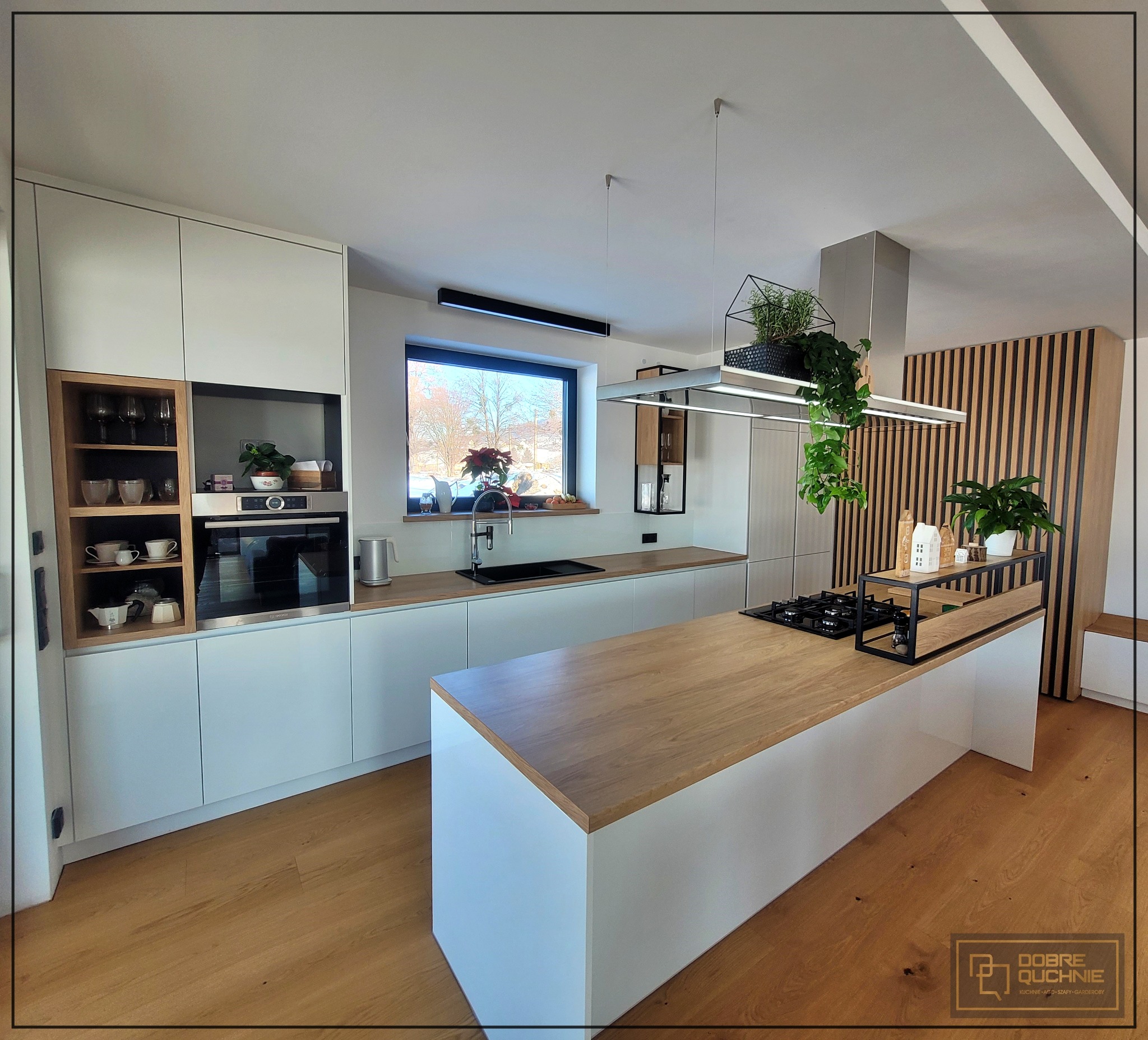 Nowoczesna kuchnia z białymi szafkami i drewnianym blatem, wyspa kuchenna z płytą grzewczą, wiszące rośliny, drewniane lamele na ścianie.
