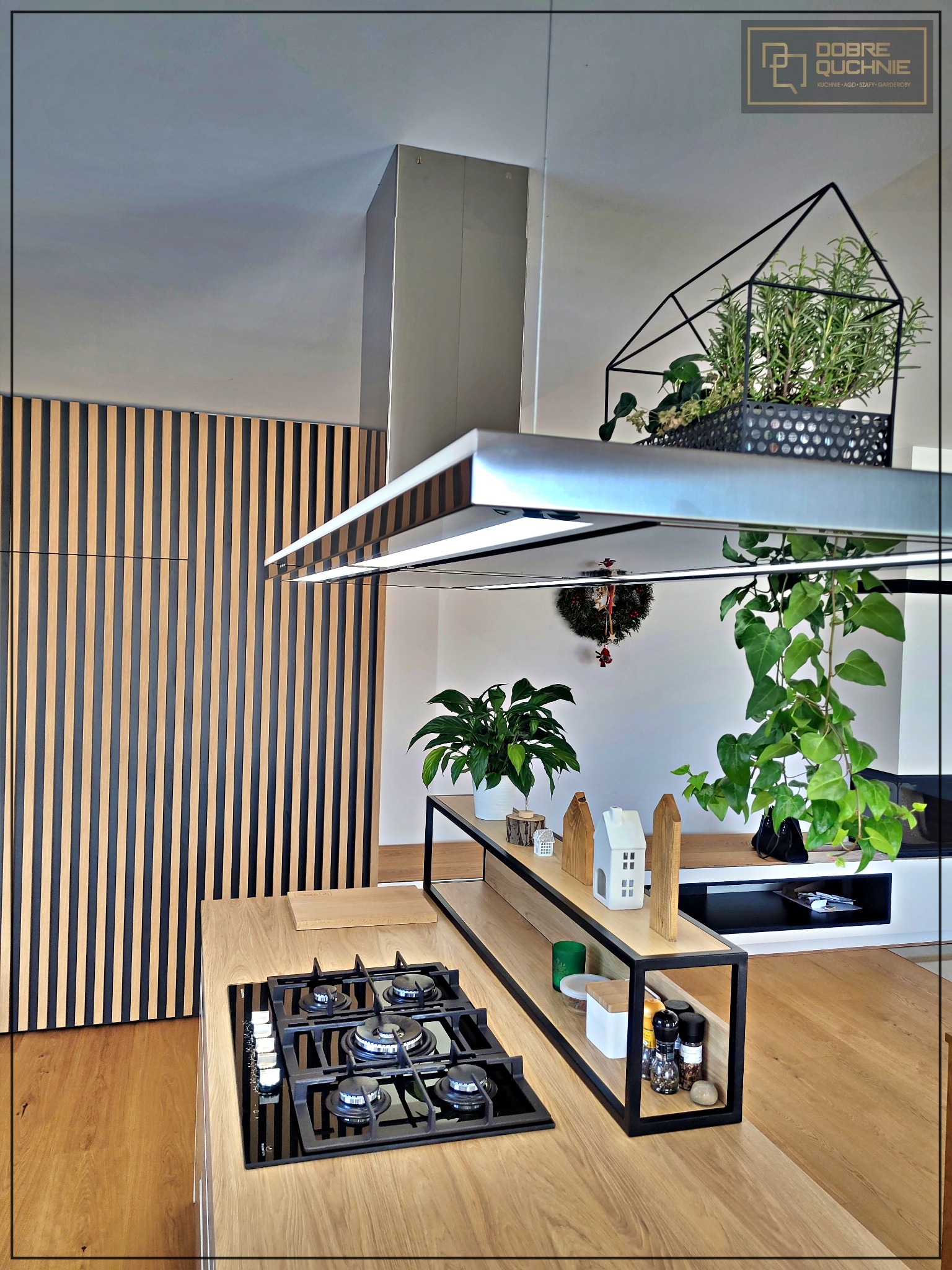 Nowoczesna kuchnia z wyspą i płytą gazową, drewniane lamele na ścianie, minimalistyczny okap, półki z dekoracjami i roślinami doniczkowymi.