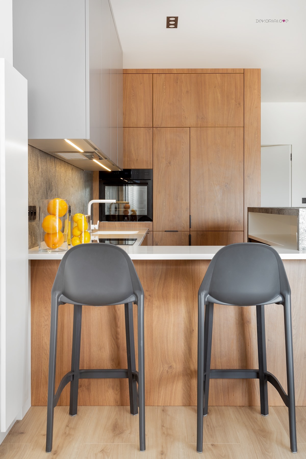 Nowoczesna kuchnia z drewnianymi frontami, białym blatem i dwoma szarymi stołkami barowymi; na blacie szklane naczynia z pomarańczami i cytrynami.