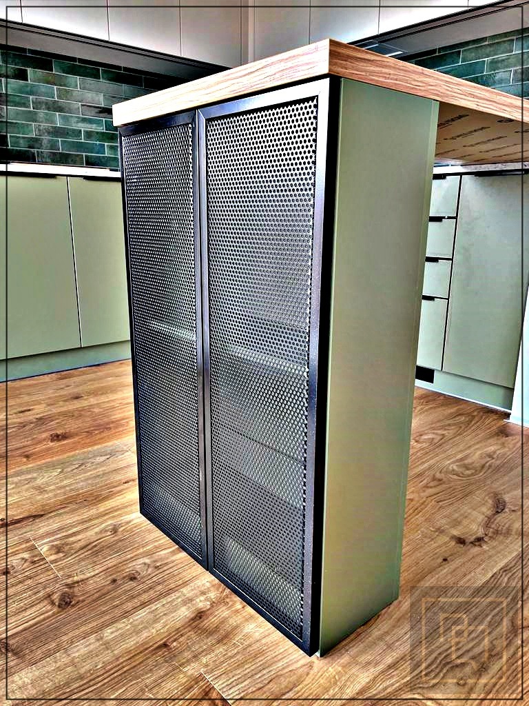 Wyspa kuchenna z metalowymi drzwiczkami z perforowanej blachy, drewniany blat, zielone szafki w tle i podłoga z paneli imitujących drewno.