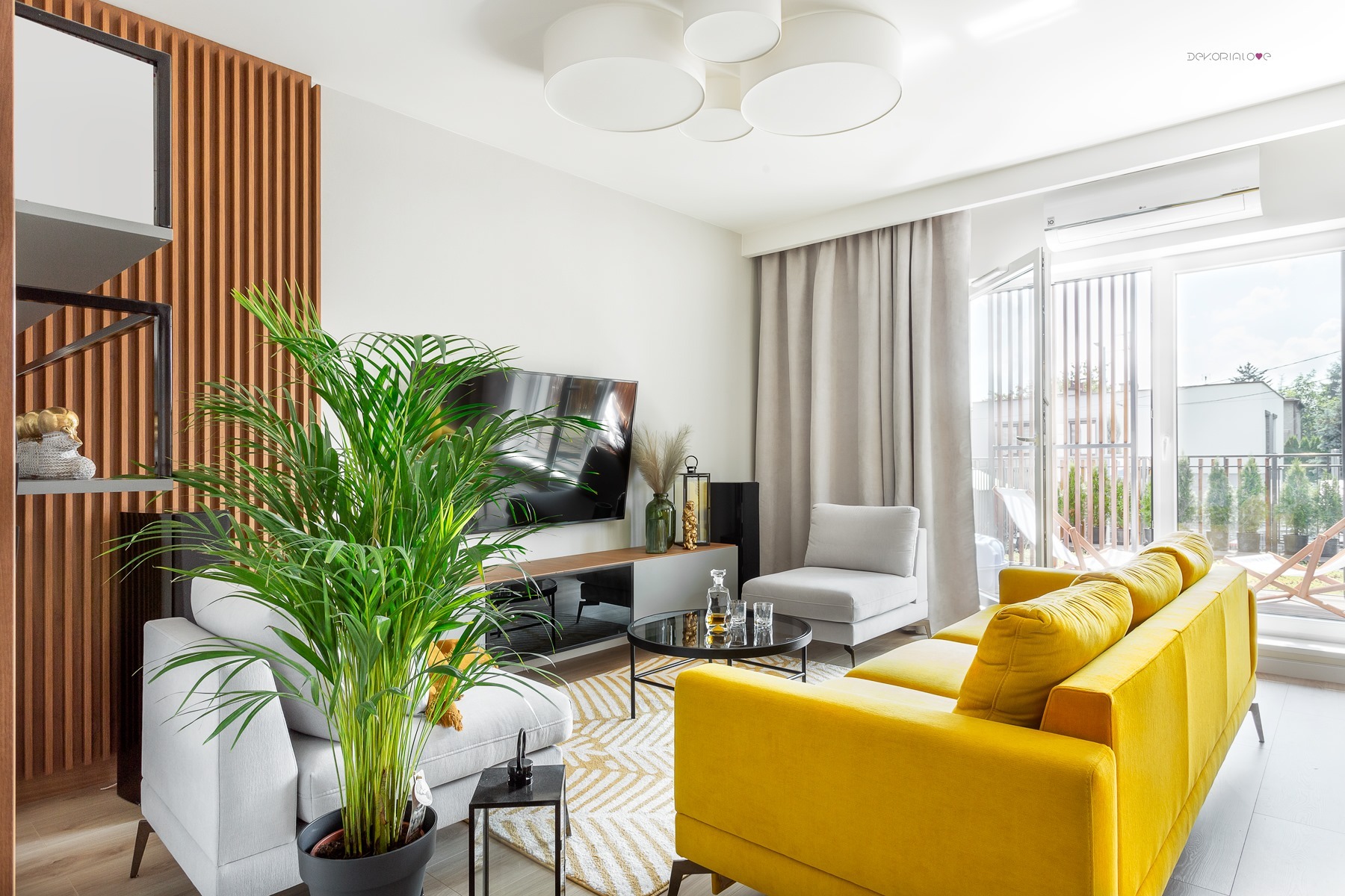 Jasne, przestronne wnętrze salonu z żółtą sofą, szarymi fotelami, telewizorem i drewnianą ścianką działową. Na pierwszym planie duża roślina doniczkowa. Widok na balkon przez otwarte drzwi.