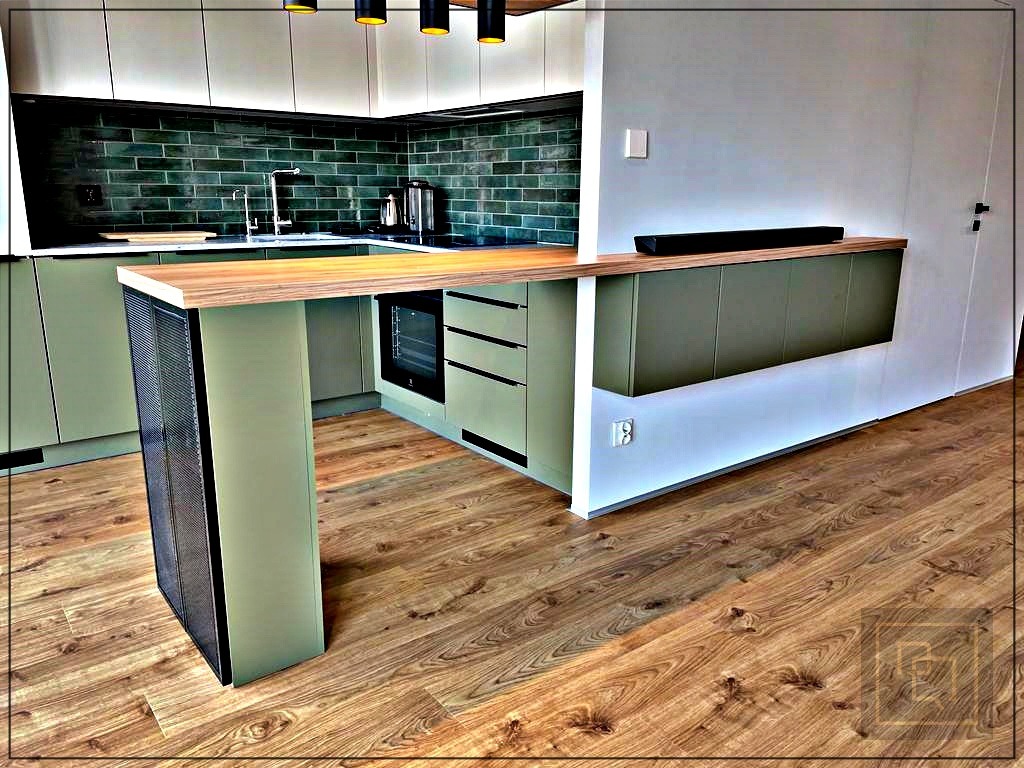 Nowoczesna kuchnia z zielonymi frontami, drewnianym blatem i wyspą z metalową kratką, oświetlenie punktowe, ciemnozielona glazura na ścianie.