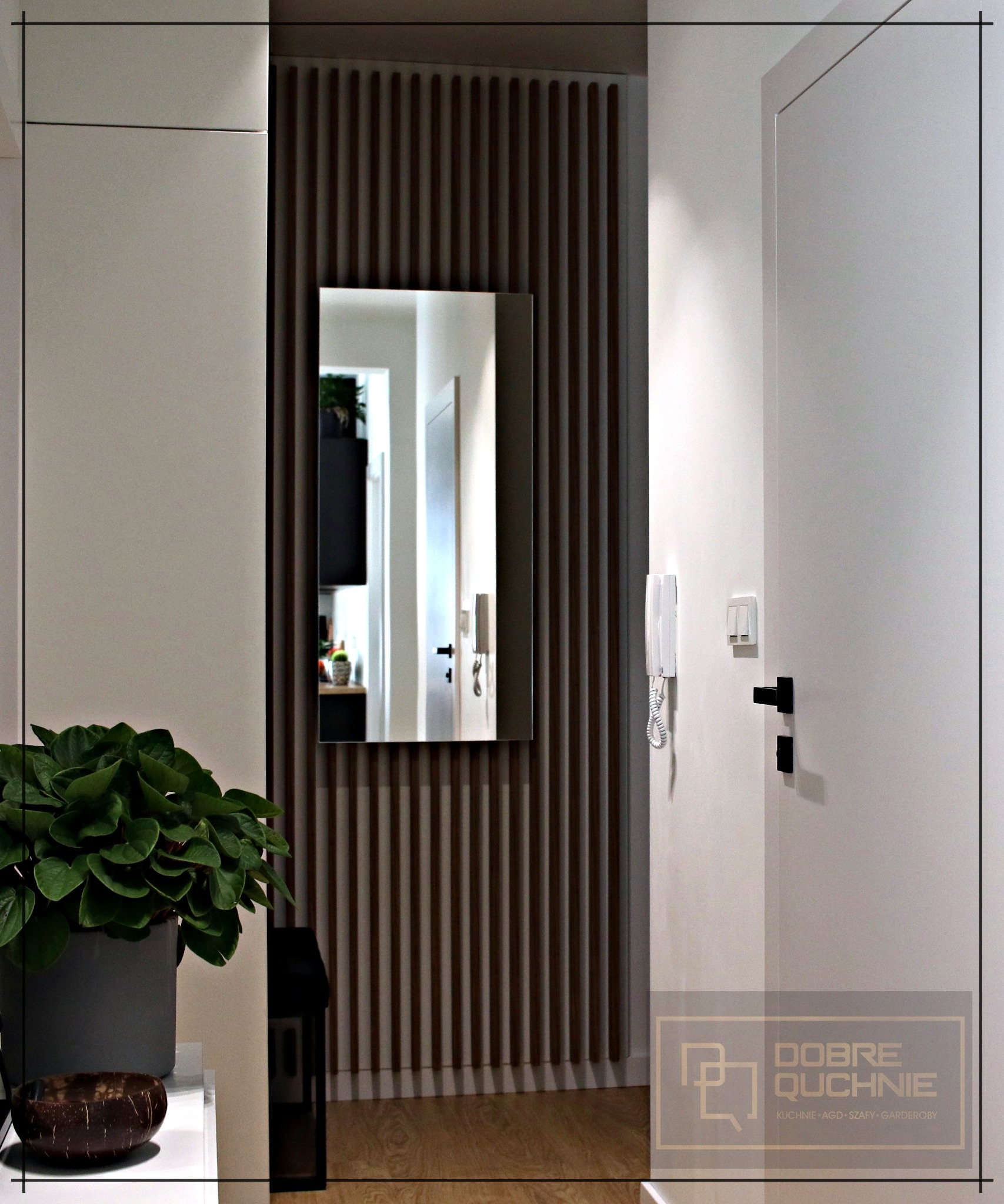 Nowoczesny przedpokój z lustrem w wąskiej ramie na tle drewnianej ściany z pionowych listewek, obok białe drzwi i interkom, w tle widoczna kuchnia.