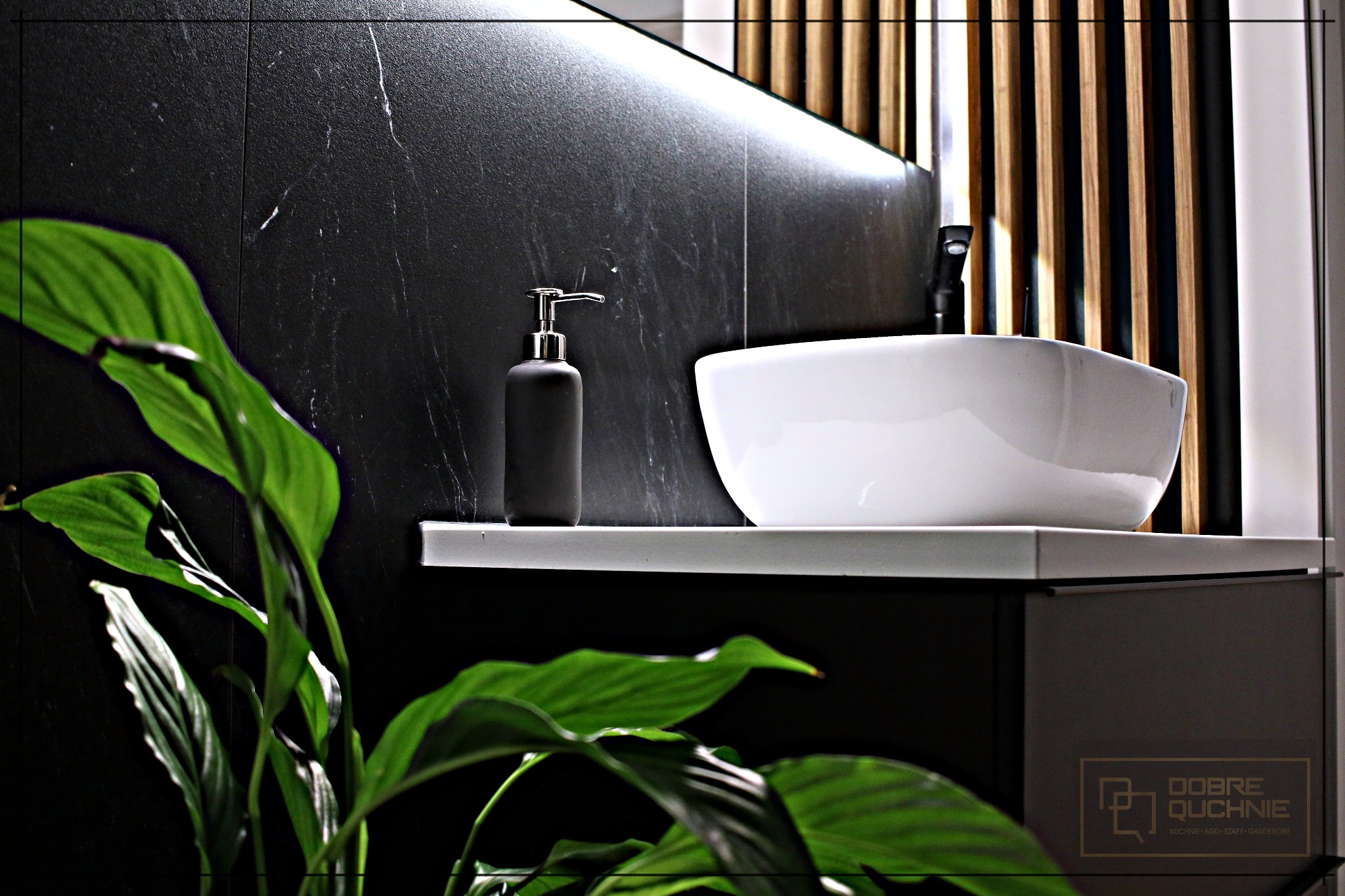Nowoczesna łazienka z umywalką nablatową, czarną marmurową ścianą i rośliną doniczkową w pierwszym planie, z widocznym dozownikiem mydła w kolorze czarnym.