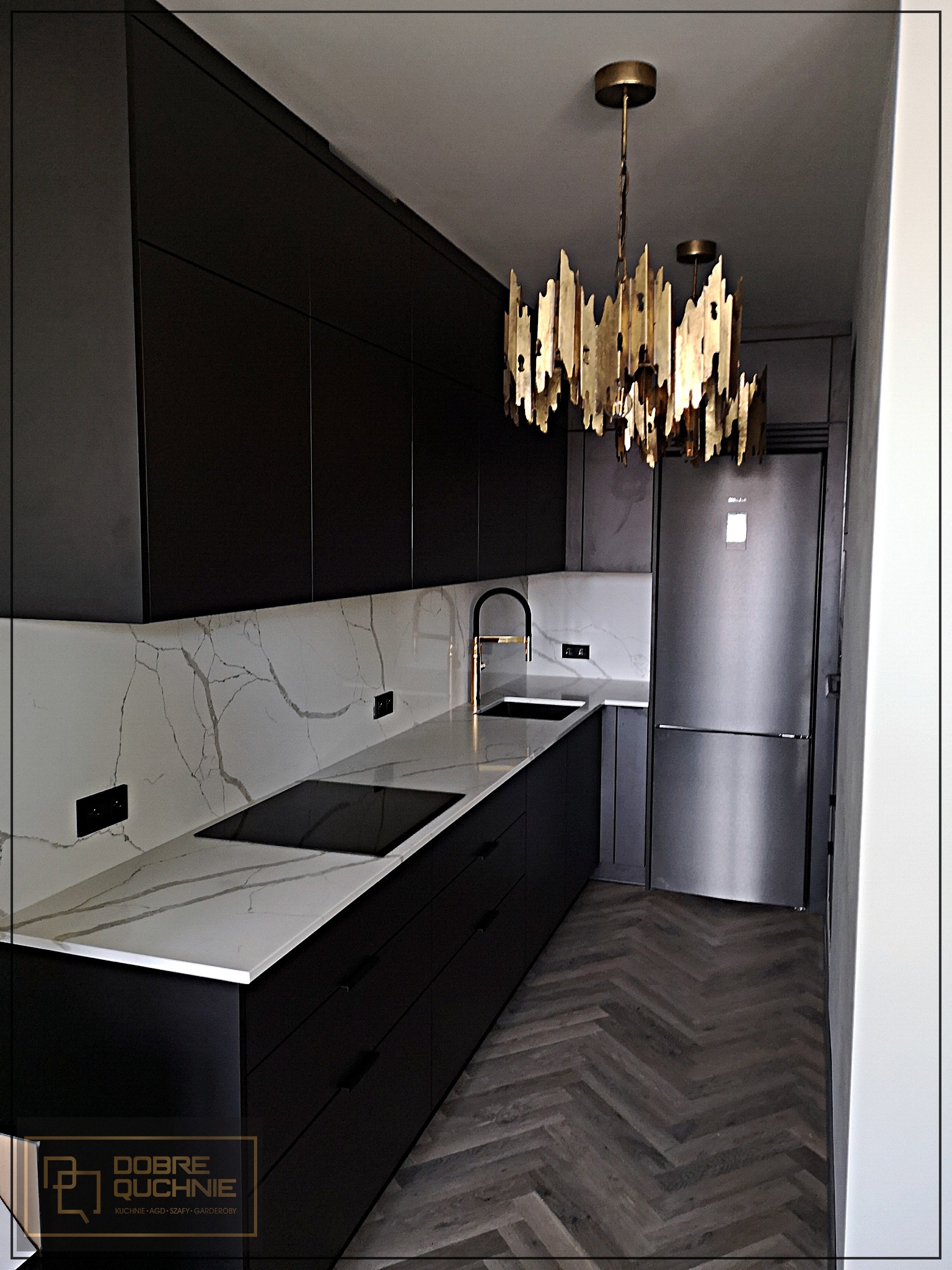 Nowoczesna kuchnia z czarnymi matowymi szafkami, blatem z białego marmuru z szarymi żyłami, złotą armaturą i designerską lampą w stylu art deco, parkiet w jodełkę.