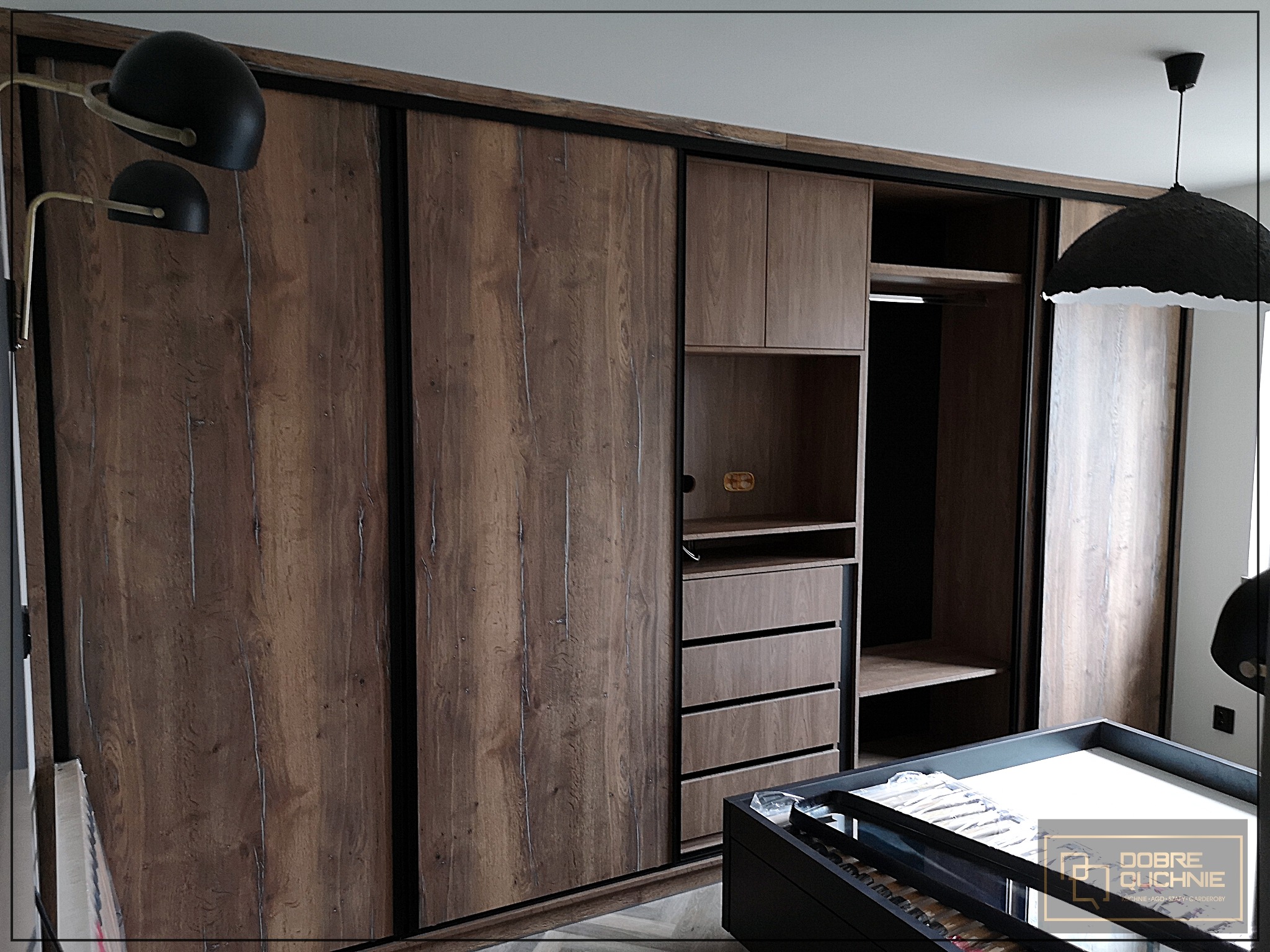 Szafa wnękowa z przesuwnymi drzwiami w odcieniach brązu i czerni, z widocznymi półkami i szufladami, oświetlona lampą wiszącą i kinkietem, z logo firmy w prawym dolnym rogu.