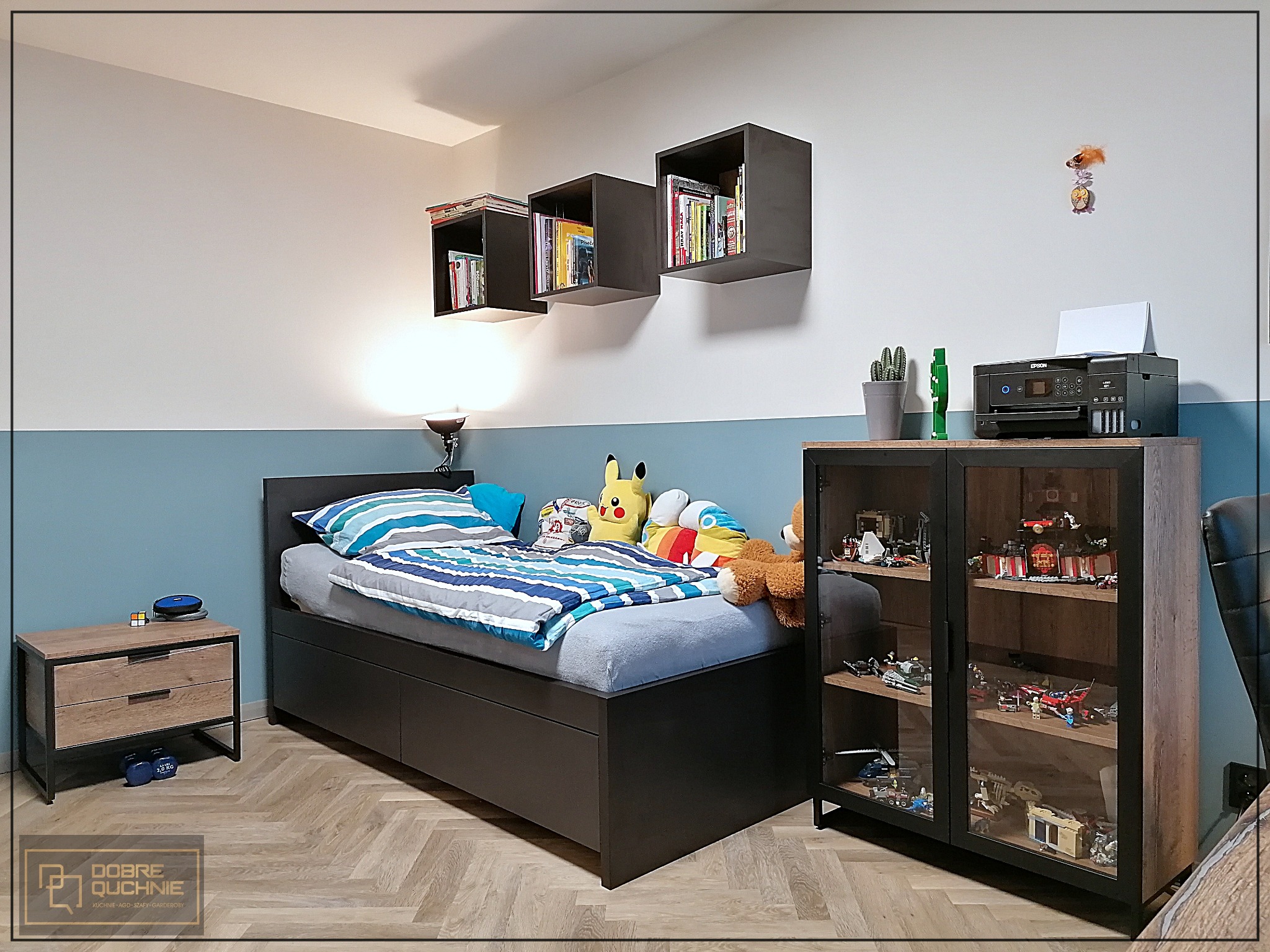 Pokój dziecięcy z łóżkiem z szufladami, szafką nocną z dwiema szufladami, wiszącymi półkami w formie kostek i przeszkloną szafką z kolekcją klocków Lego.