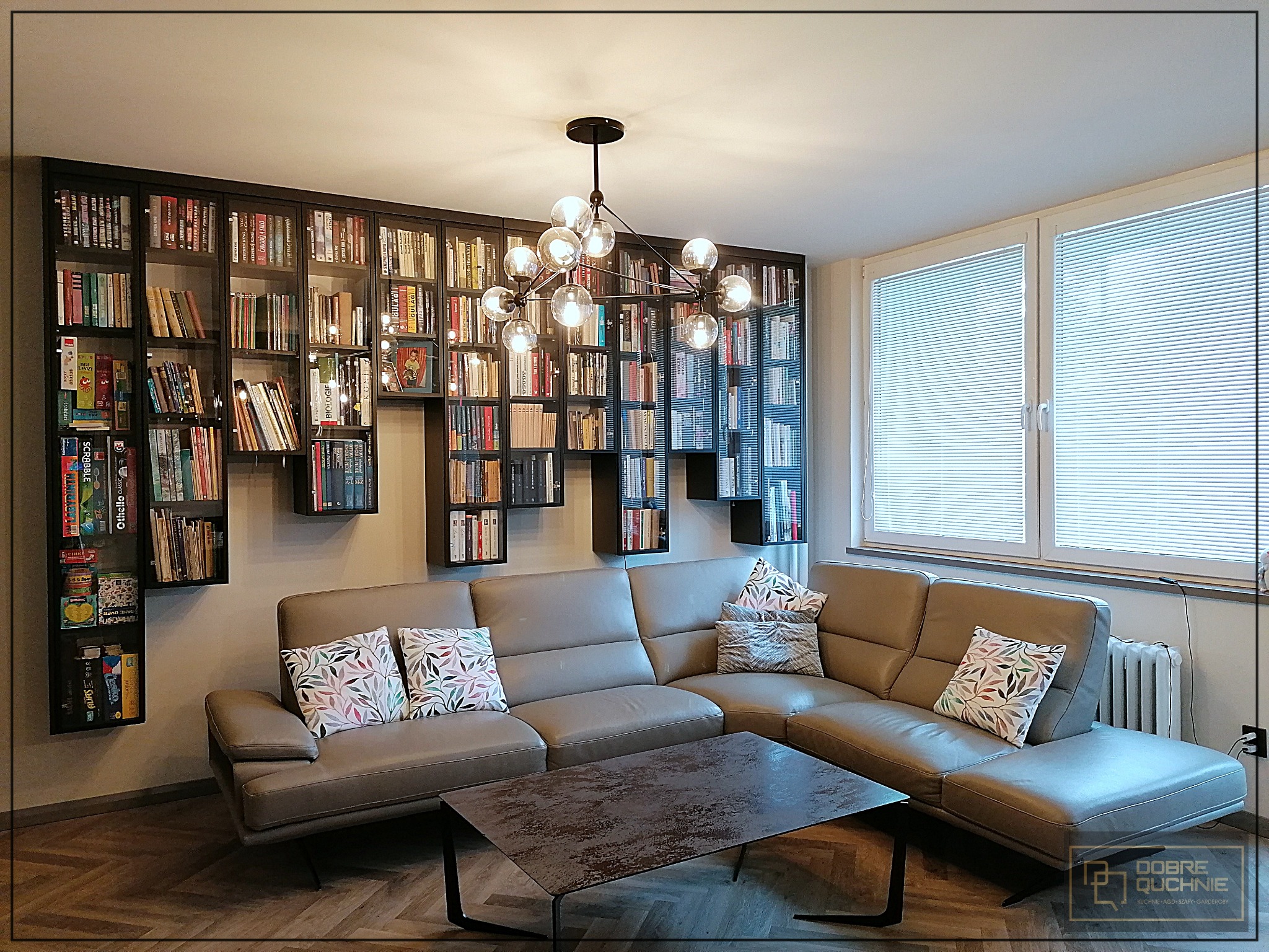 Salon z szarą sofą narożną, stolikiem kawowym i regałem na książki w zabudowie, oświetlonym nowoczesną lampą wiszącą z żarówkami kulistymi.