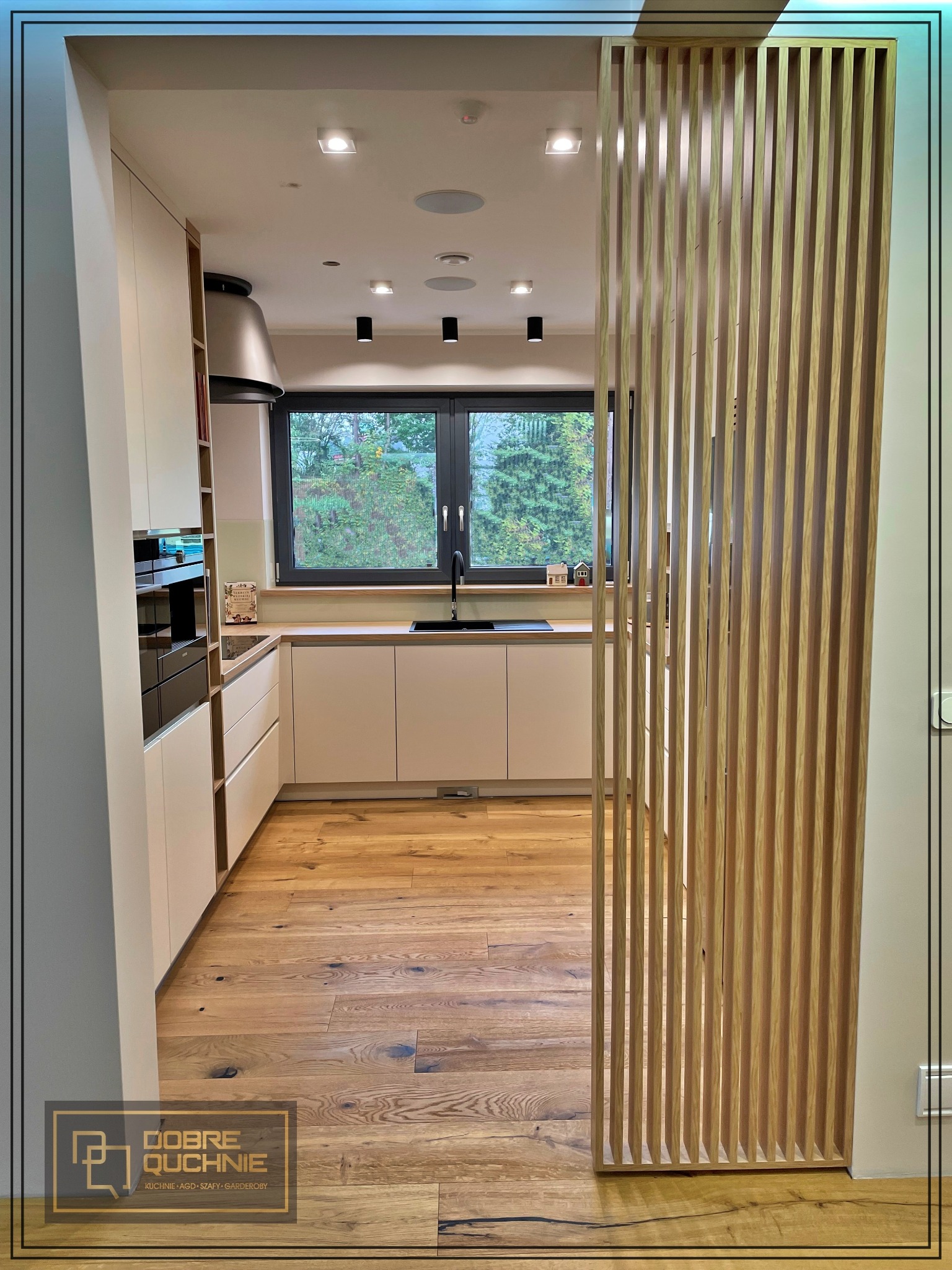 Jasna kuchnia z drewnianą podłogą i lamelową ścianką działową, widok na okno z zielenią na zewnątrz, nowoczesny design.