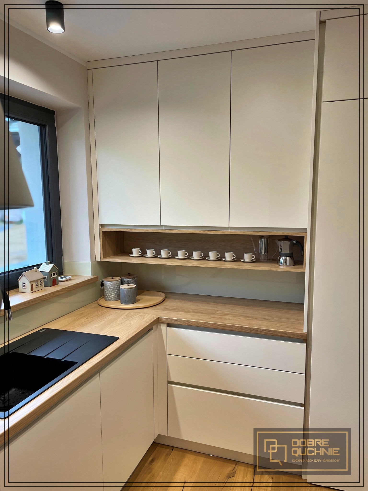 Jasna kuchnia na wymiar w Cieszynie z drewnianym blatem, czarnym zlewem i rzędem filiżanek na półce, uzupełniona o ekspres do kawy.