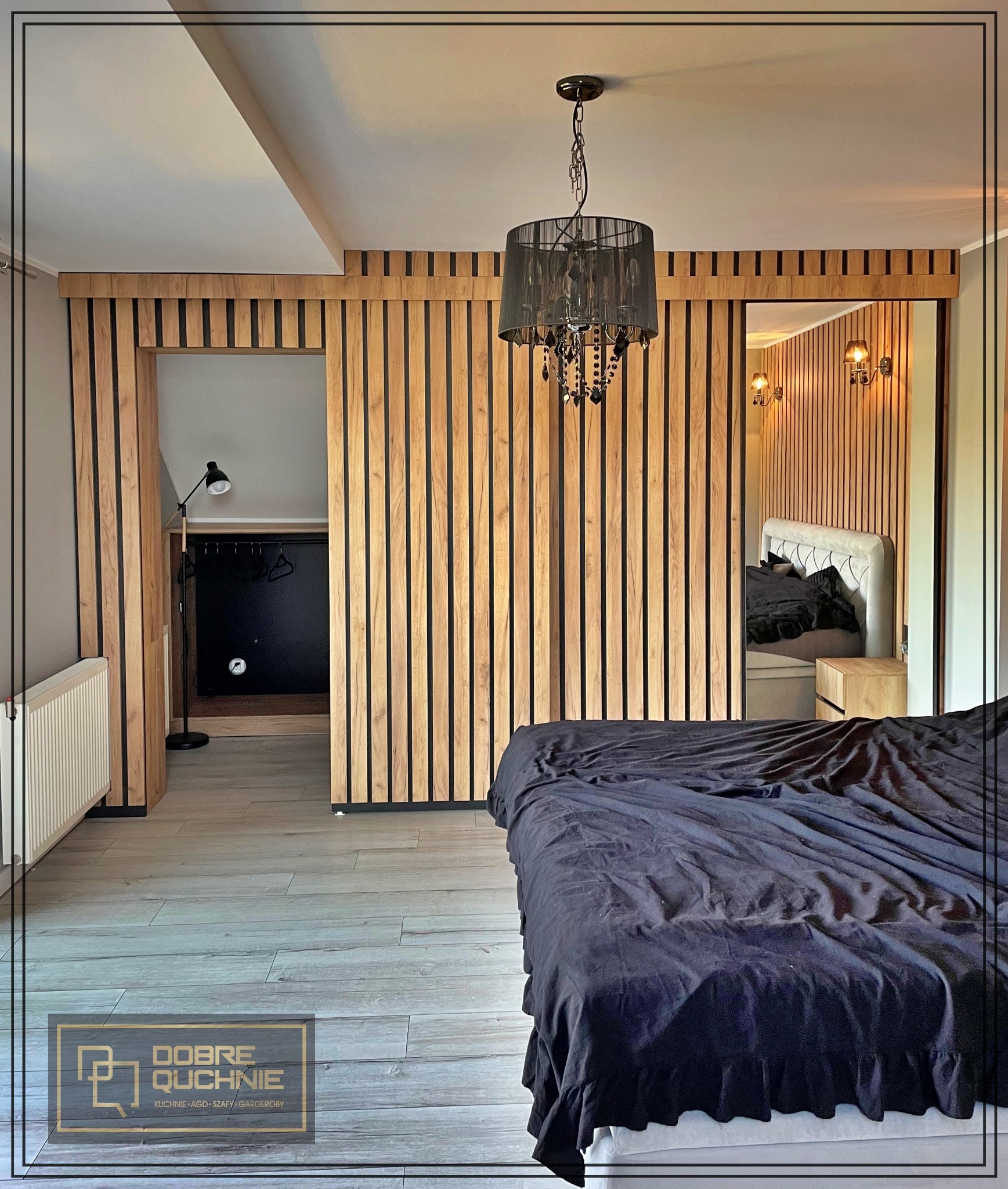 Sypialnia z drewnianą zabudową lamelową, czarnym żyrandolem i wejściem do garderoby z czarną ścianą i lampą podłogową. W lustrze odbicie łóżka z beżowym zagłówkiem i drewnianą szafką nocną.